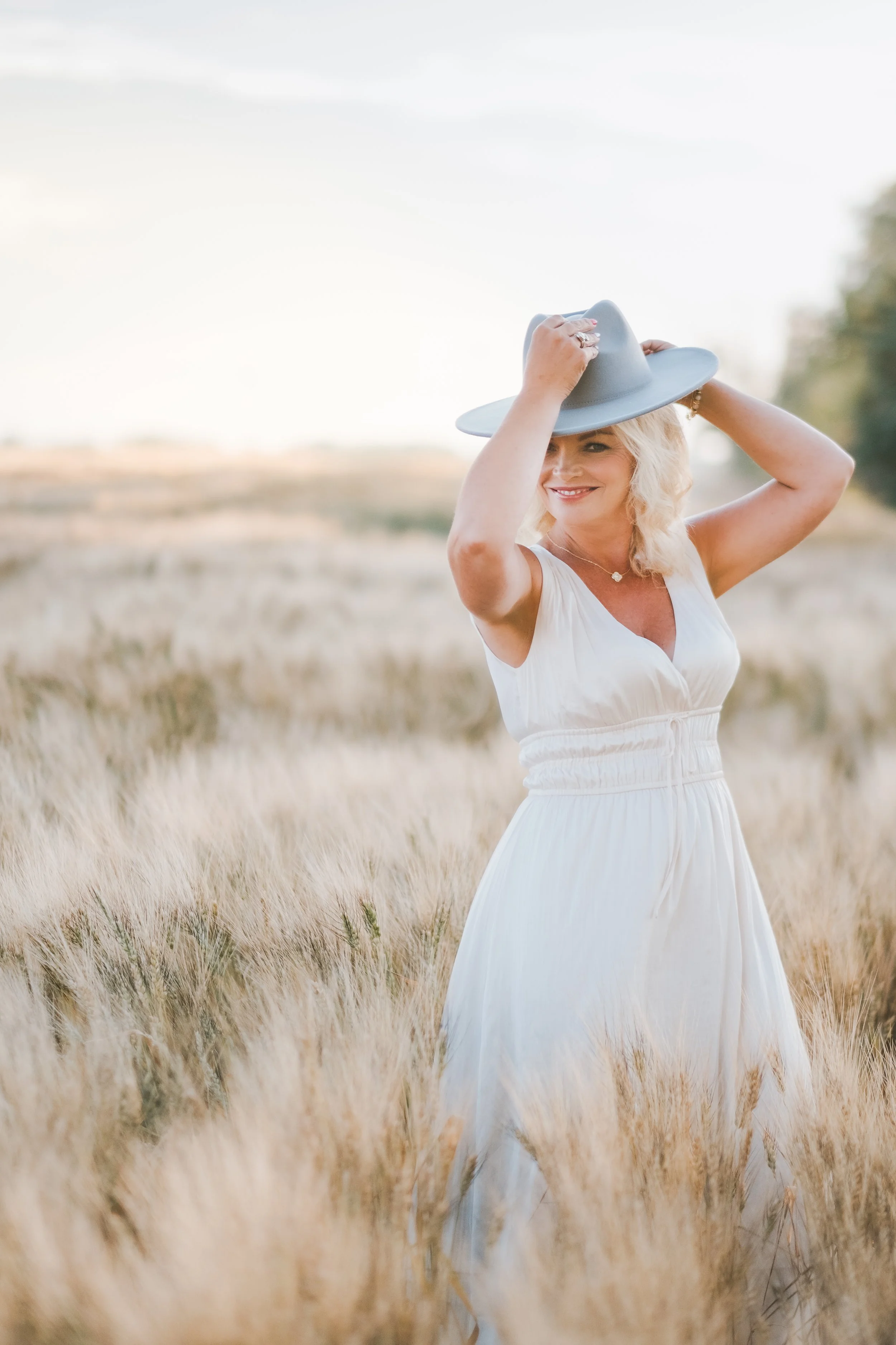 A smiling woman in a white dress standing in a field of tall grass, holding a large gray hat on her head.