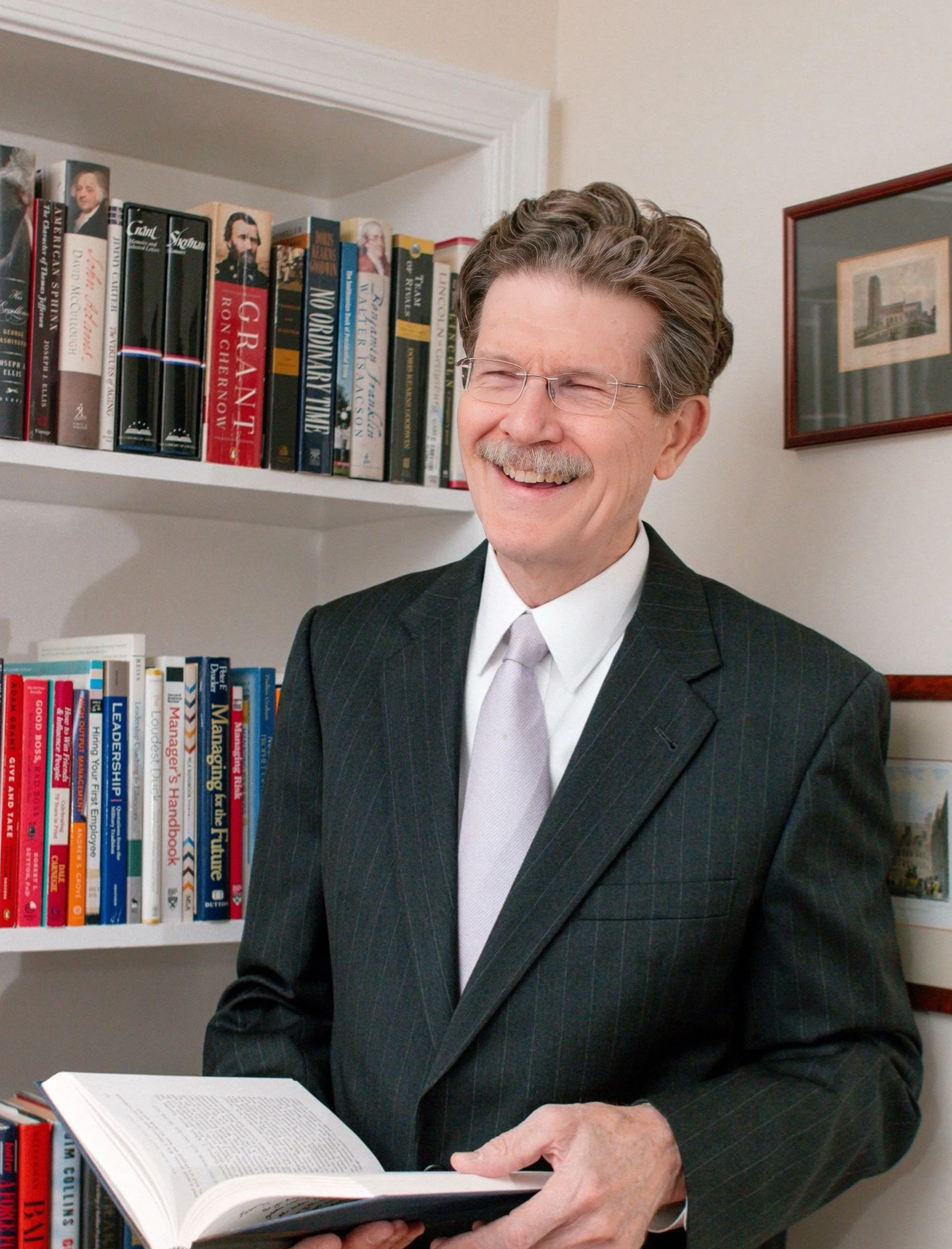 A smiling man with glasses and a gray mustache, wearing a dark pinstripe suit and a light-colored tie, holding an open book in a room with bookshelves and framed pictures on the wall.