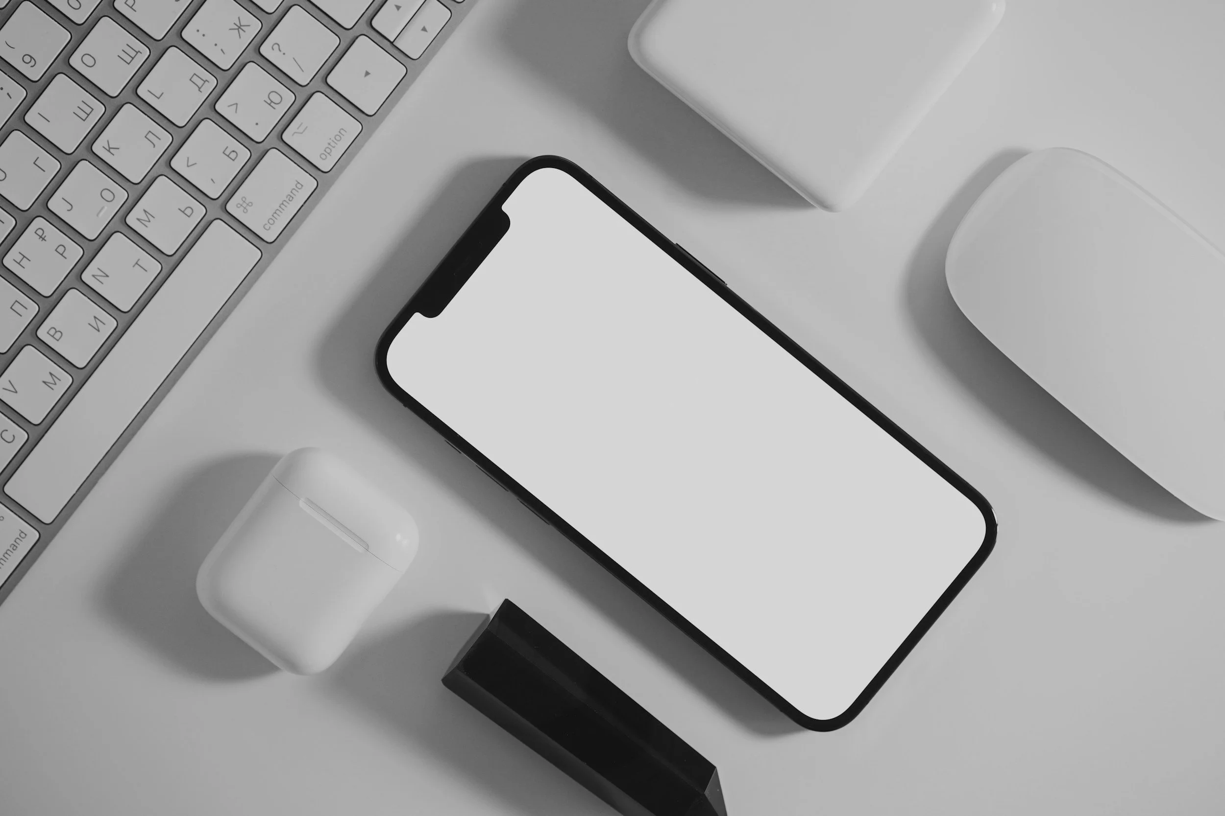 A top-down view of a minimalist workspace with a black smartphone, white wireless earbuds case, white wireless charging pad, black lipstick, and a white Apple keyboard.