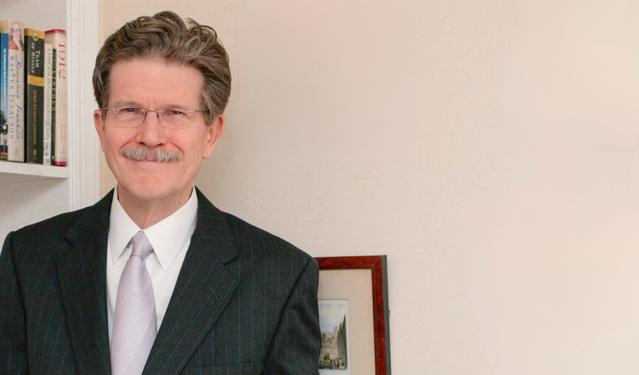 A man with glasses and a mustache dressed in a dark pinstripe suit, white shirt, and light-colored tie standing in front of a bookshelf and a framed picture on a plain wall.