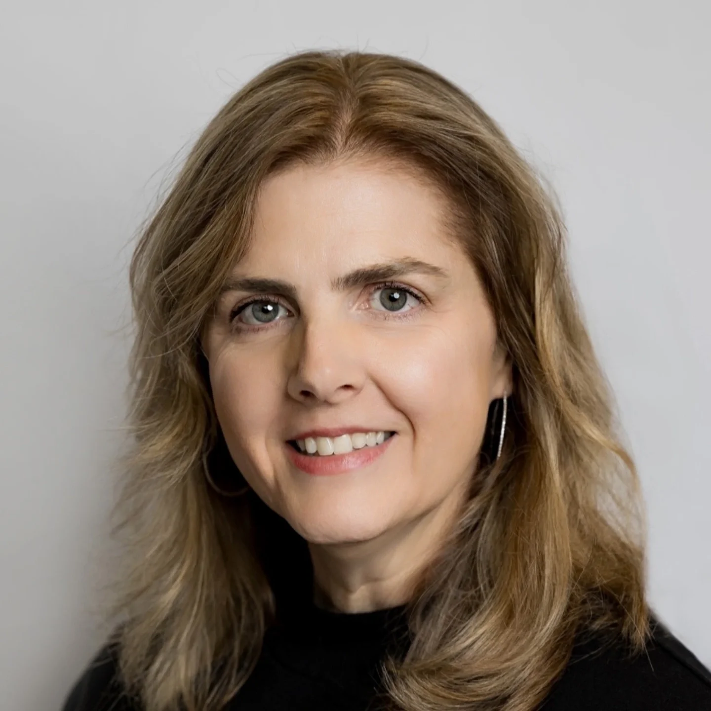 Close-up of a woman with wavy, shoulder-length brown hair, blue eyes, and light makeup, smiling slightly, wearing a black top and hoop earrings against a plain light gray background.
