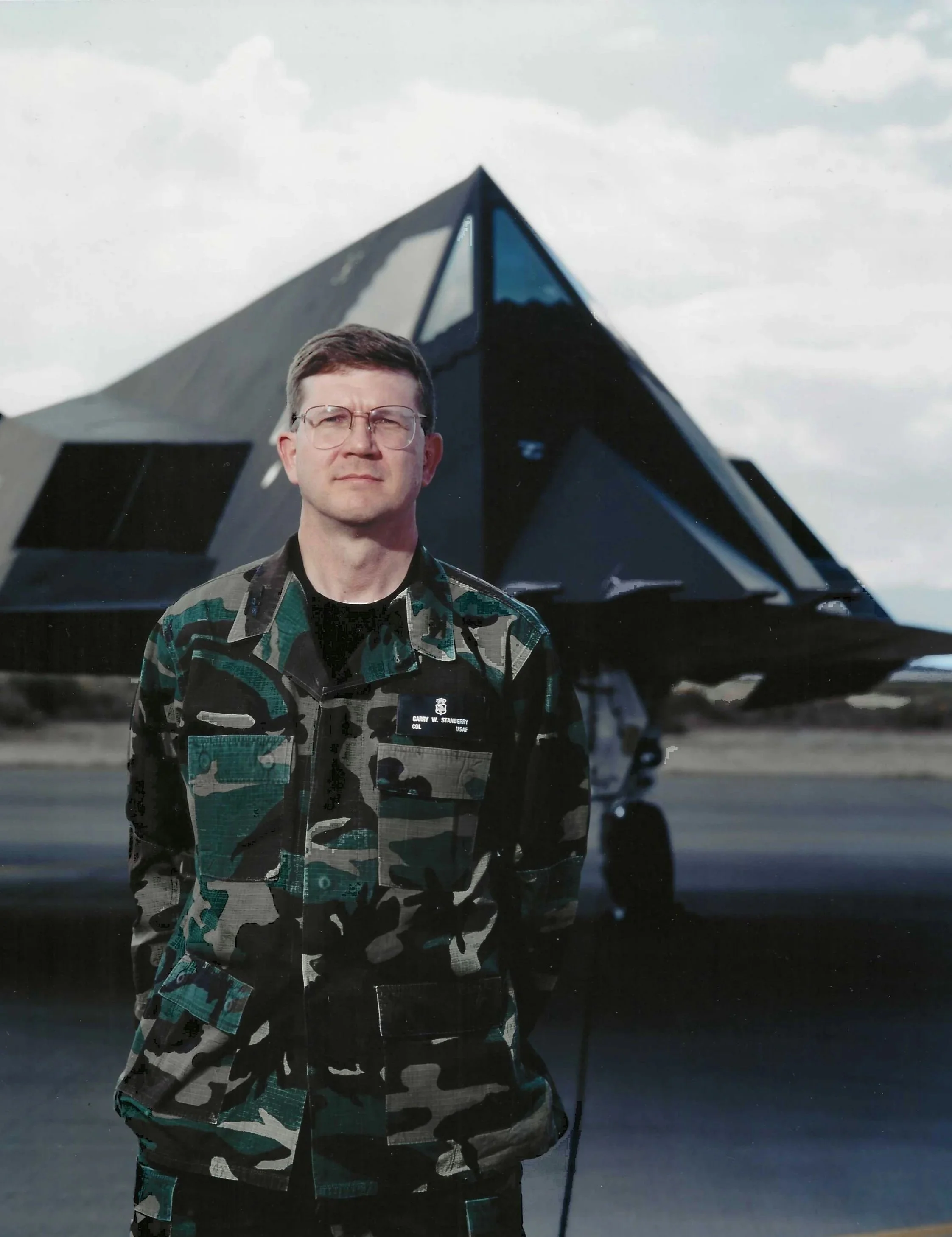 A man in military uniform standing in front of a black futuristic jet aircraft on an airfield under cloudy skies.
