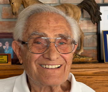 A present-day photo of Shahen Guiragossian with white hair and glasses in a cozy room with wooden walls and decorations.