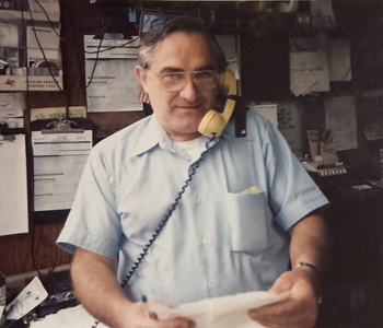 Shahen Guiragossian, dressed in a light blue shirt holding papers and talking on a yellow corded phone, in a cluttered office or workspace.