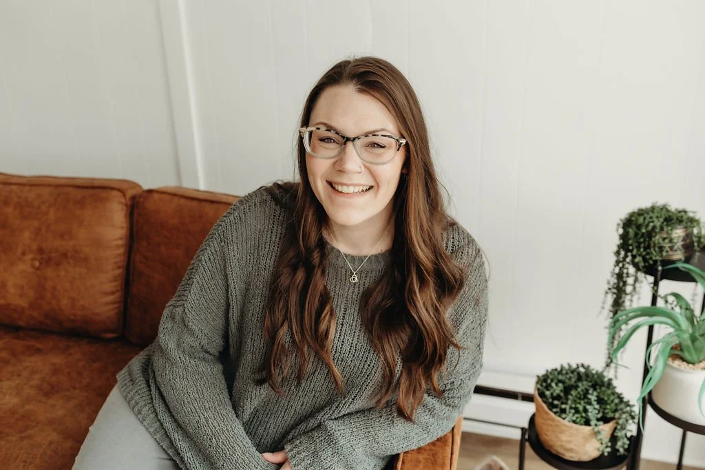 A young woman with long wavy brown hair, wearing glasses and a gray sweater, smiling and sitting on a brown couch. Therapist and counselor.