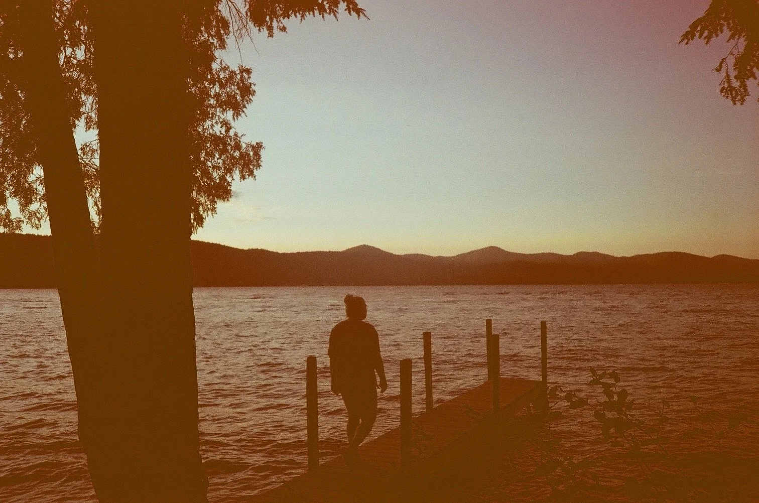 A person walking on a small dock by a lake during sunset with mountains in the background and trees framing the scene.