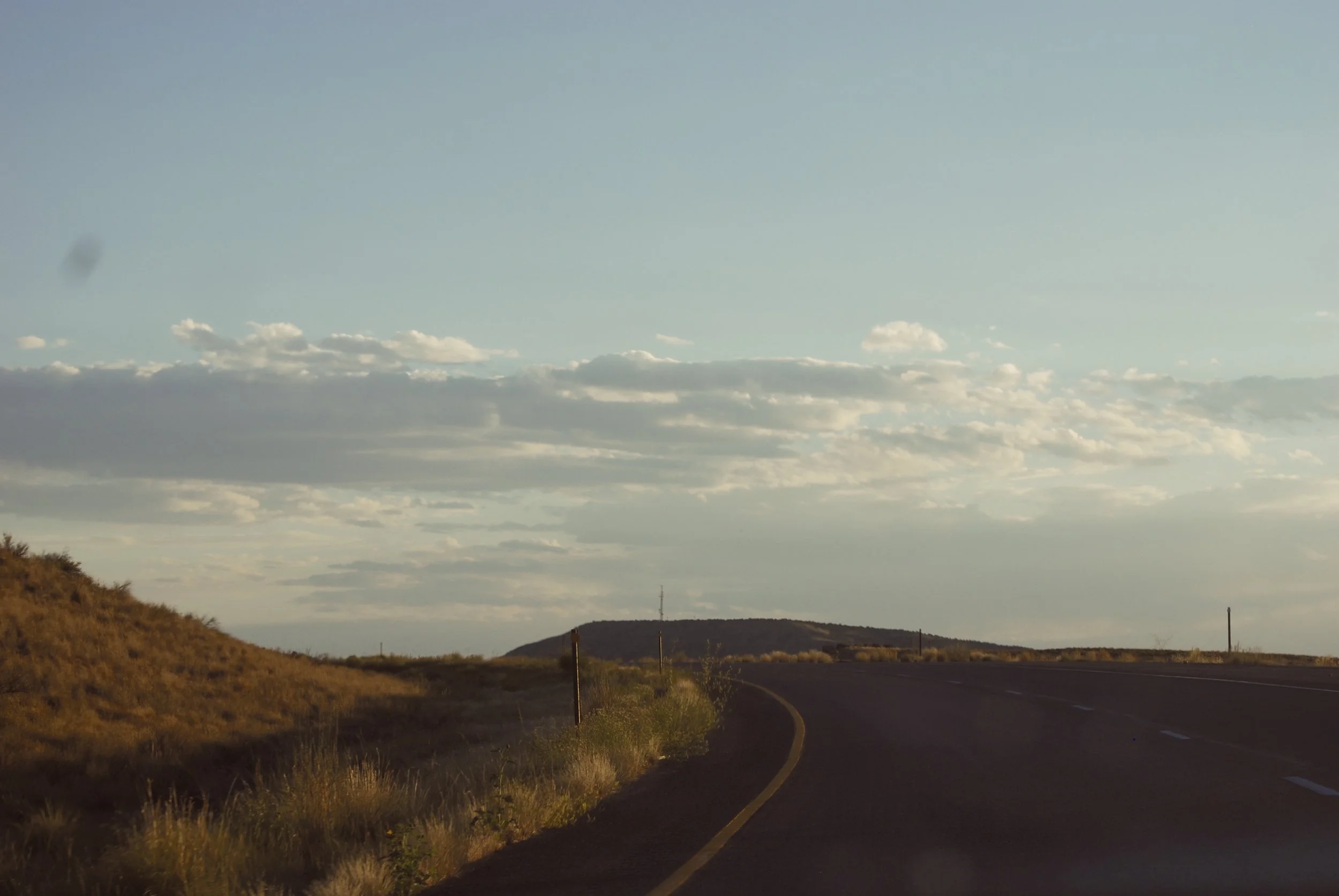 A rural highway with a curve, flanked by dry grass and a hill, under a partly cloudy sky during late afternoon.
