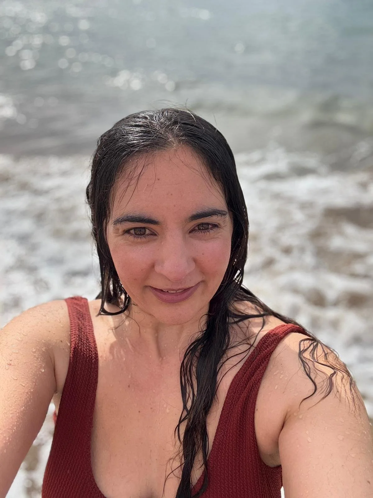 A woman with wet black hair taking a selfie at the beach, with waves in the background.