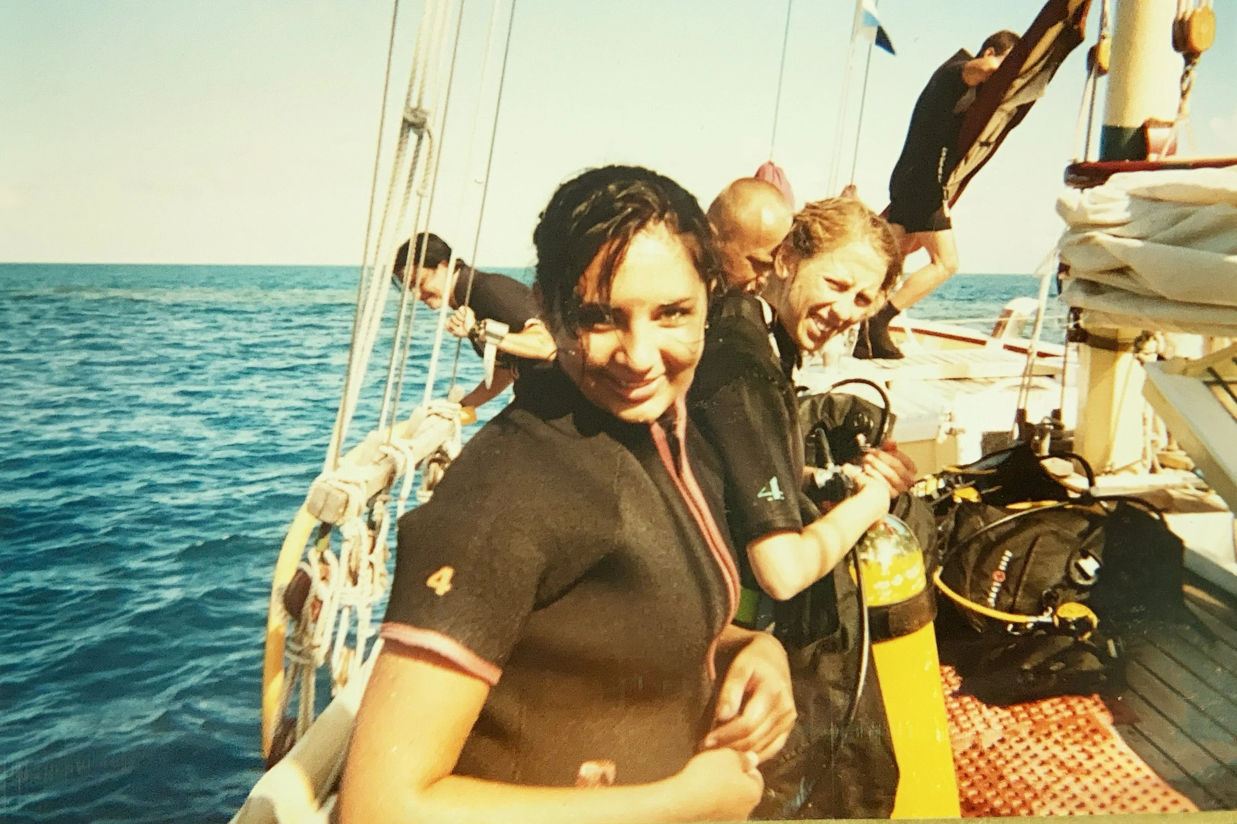 Group of people on a boat or ship, with two women smiling at the camera and others preparing gear, clouds in the sky, blue ocean in the background.