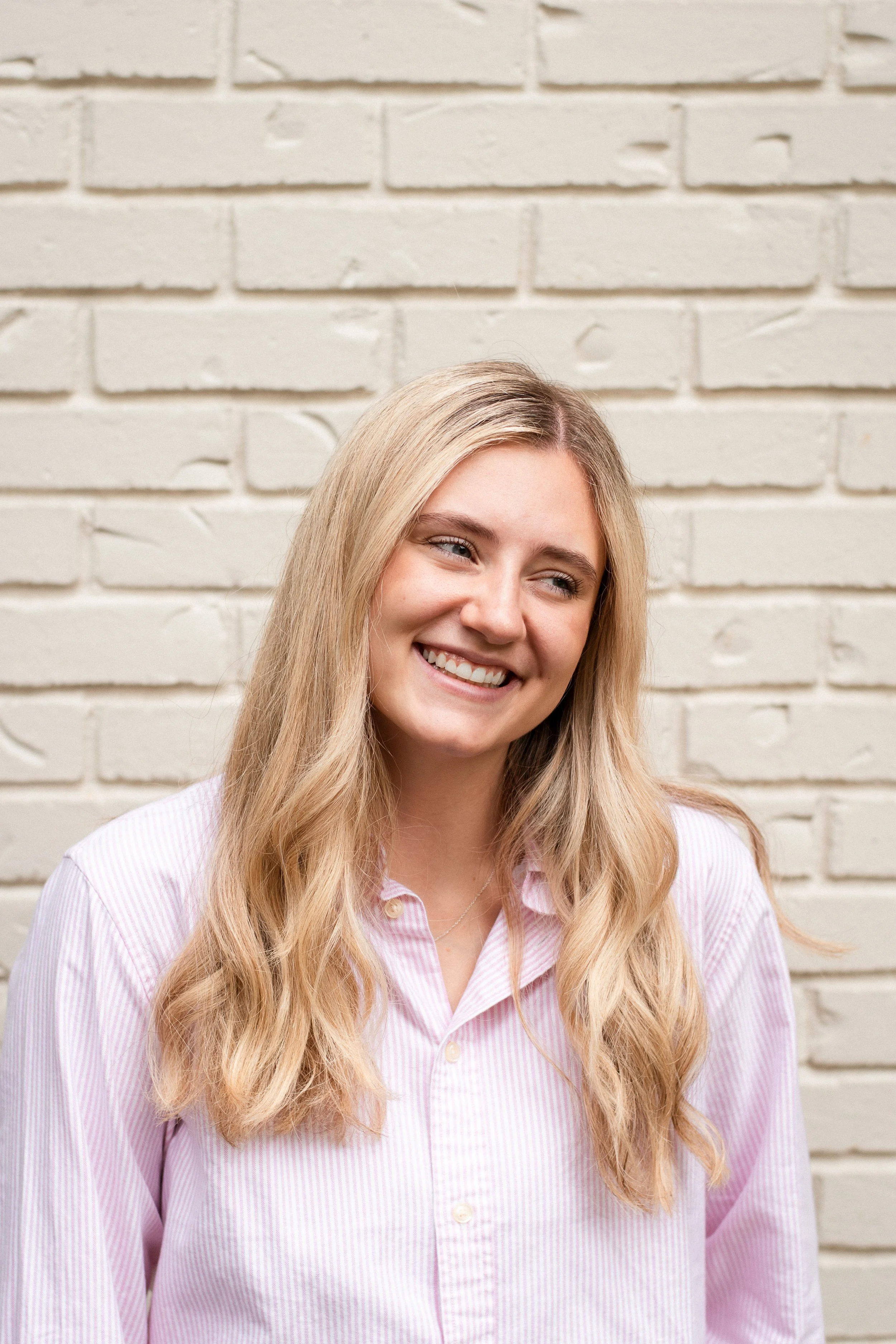 A young woman with long blonde hair smiling and looking to her right, wearing a light pink striped shirt, standing in front of a white brick wall.