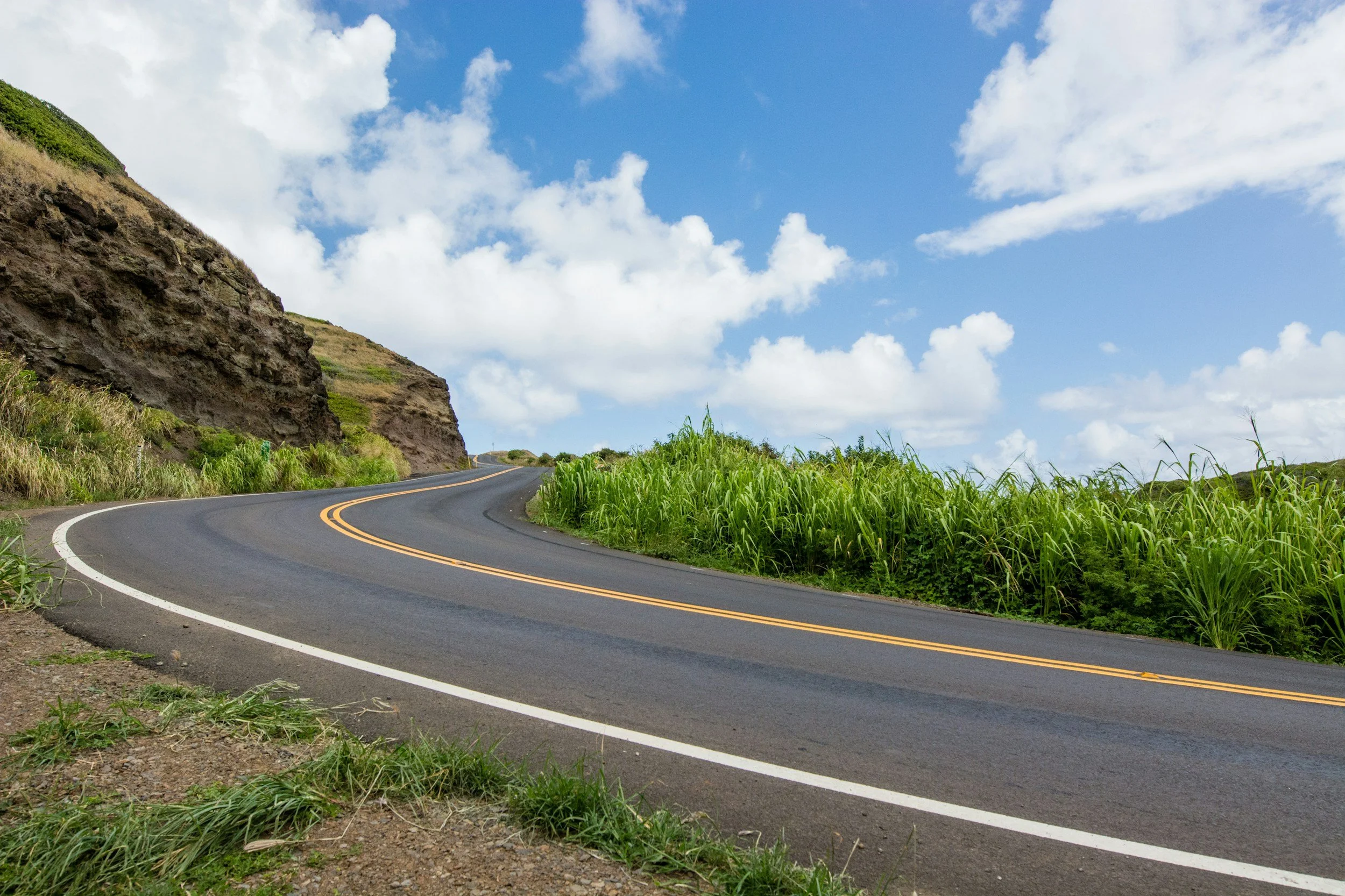 A winding paved road with double yellow lines curves past green grass and a rocky hillside under a partly cloudy blue sky.