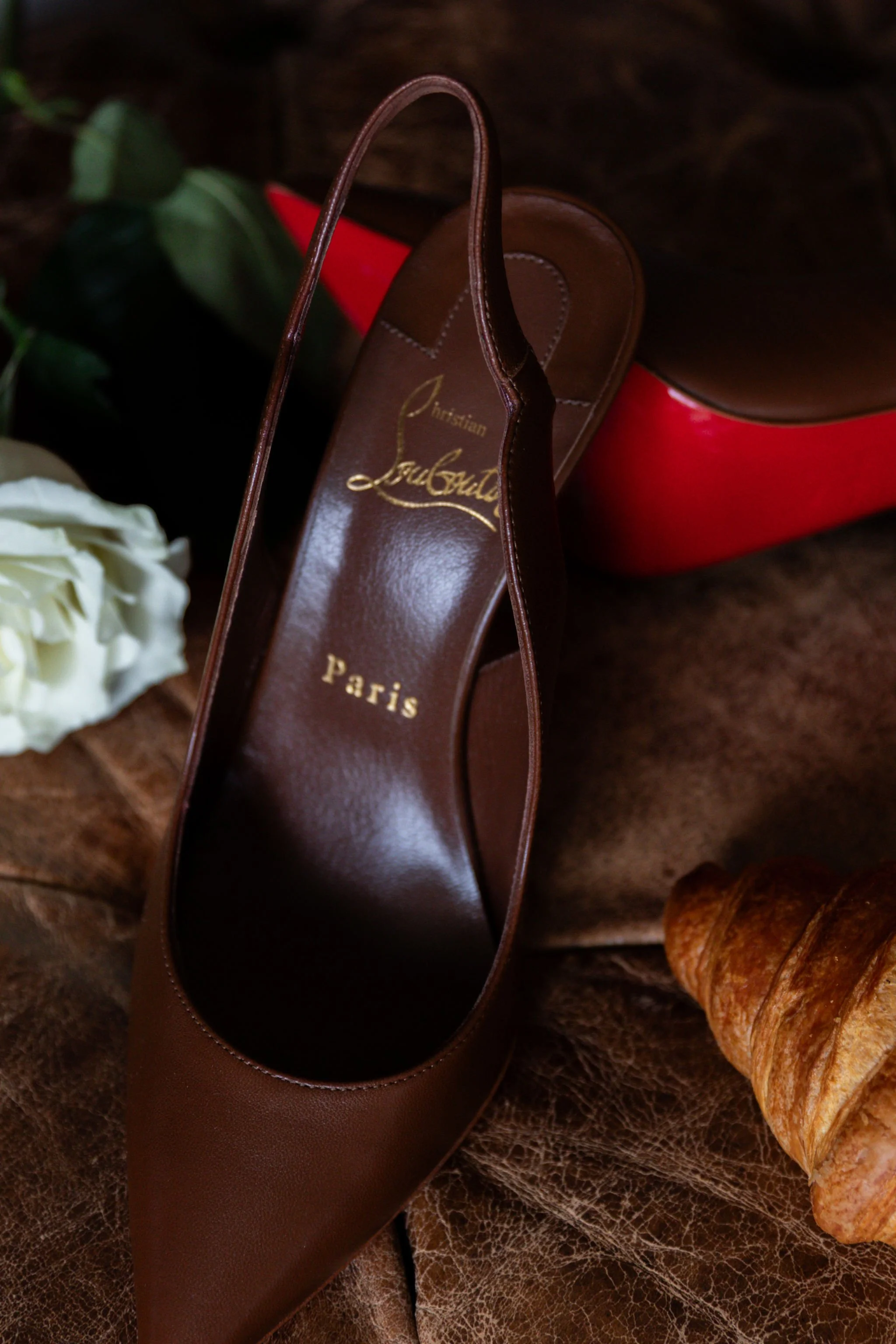 A pair of brown Christian Louboutin high heels with a red sole, a white rose, a croissant, and a red box in the background on a leather surface.