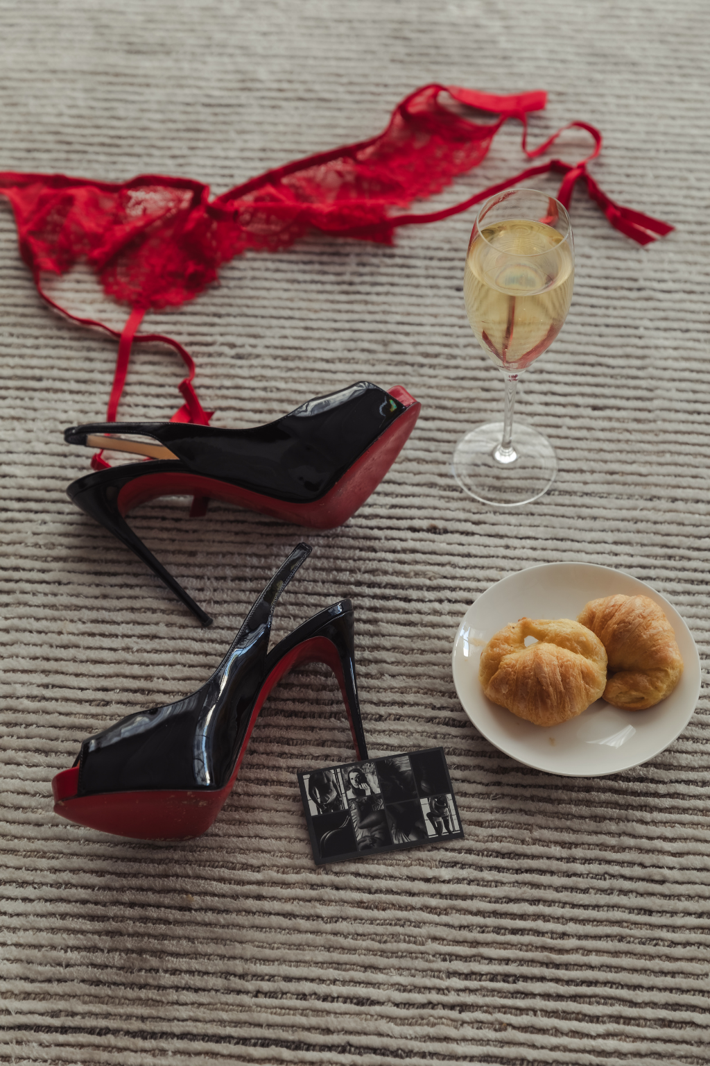 High-heeled shoes, lace lingerie, a glass of champagne, a plate with croissants, and a small photo strip on a striped carpet.