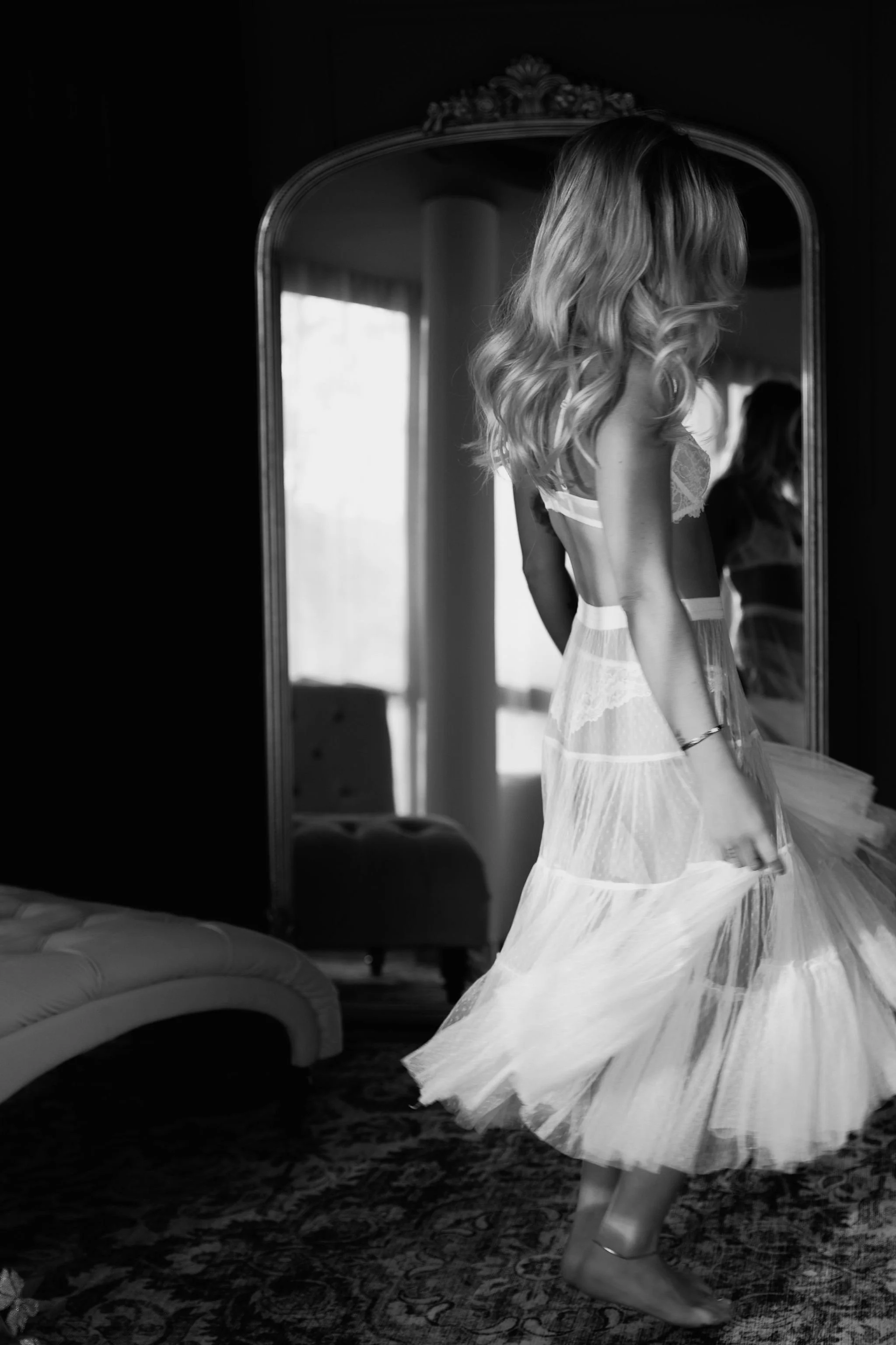 A woman with long, wavy hair wearing a light-colored lacy dress, looking in a mirror, captured in black and white.