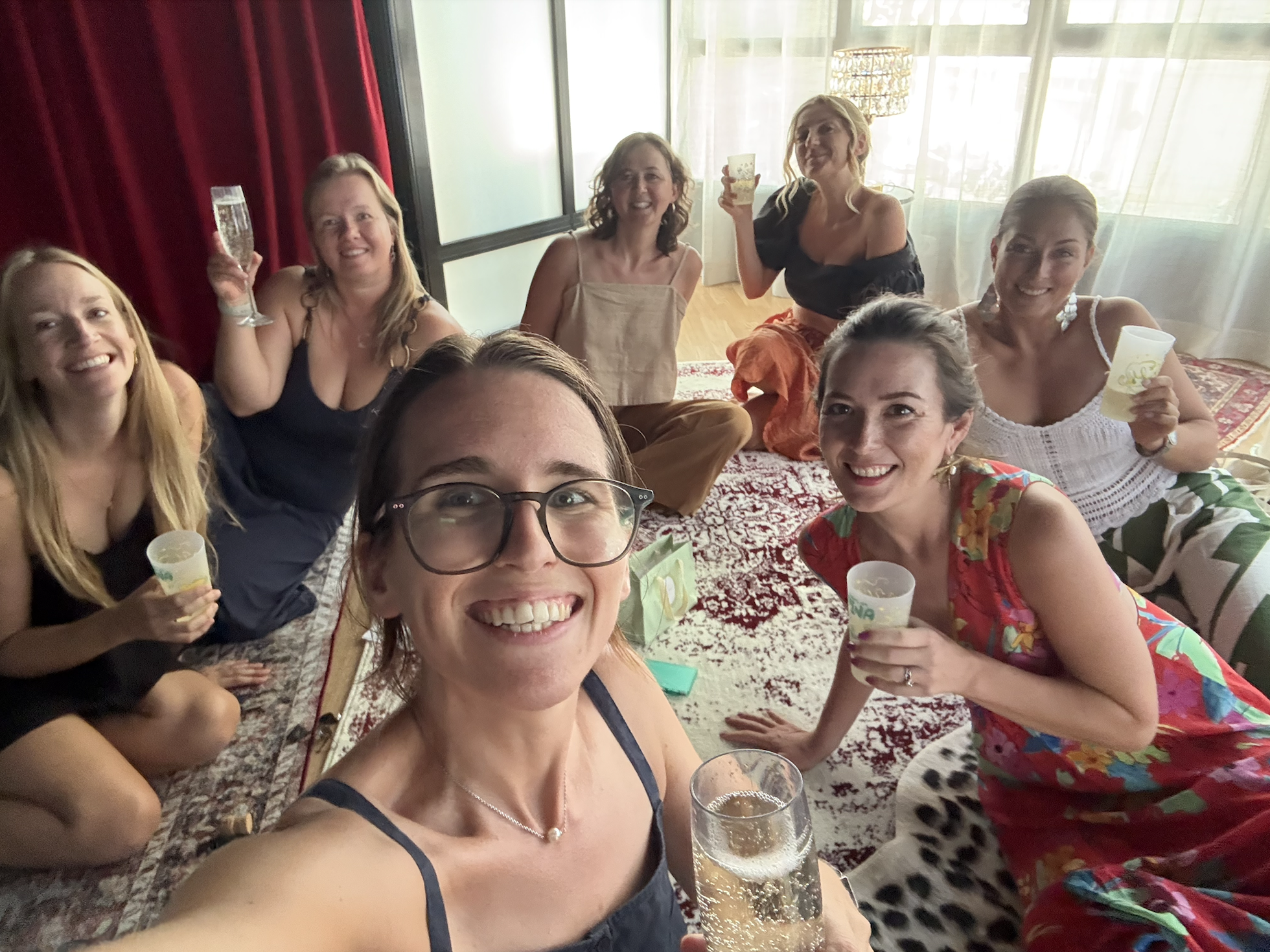 Group of women celebrating indoors, smiling, holding drinks, sitting on a decorative rug, with sunlight coming through large windows in the background.