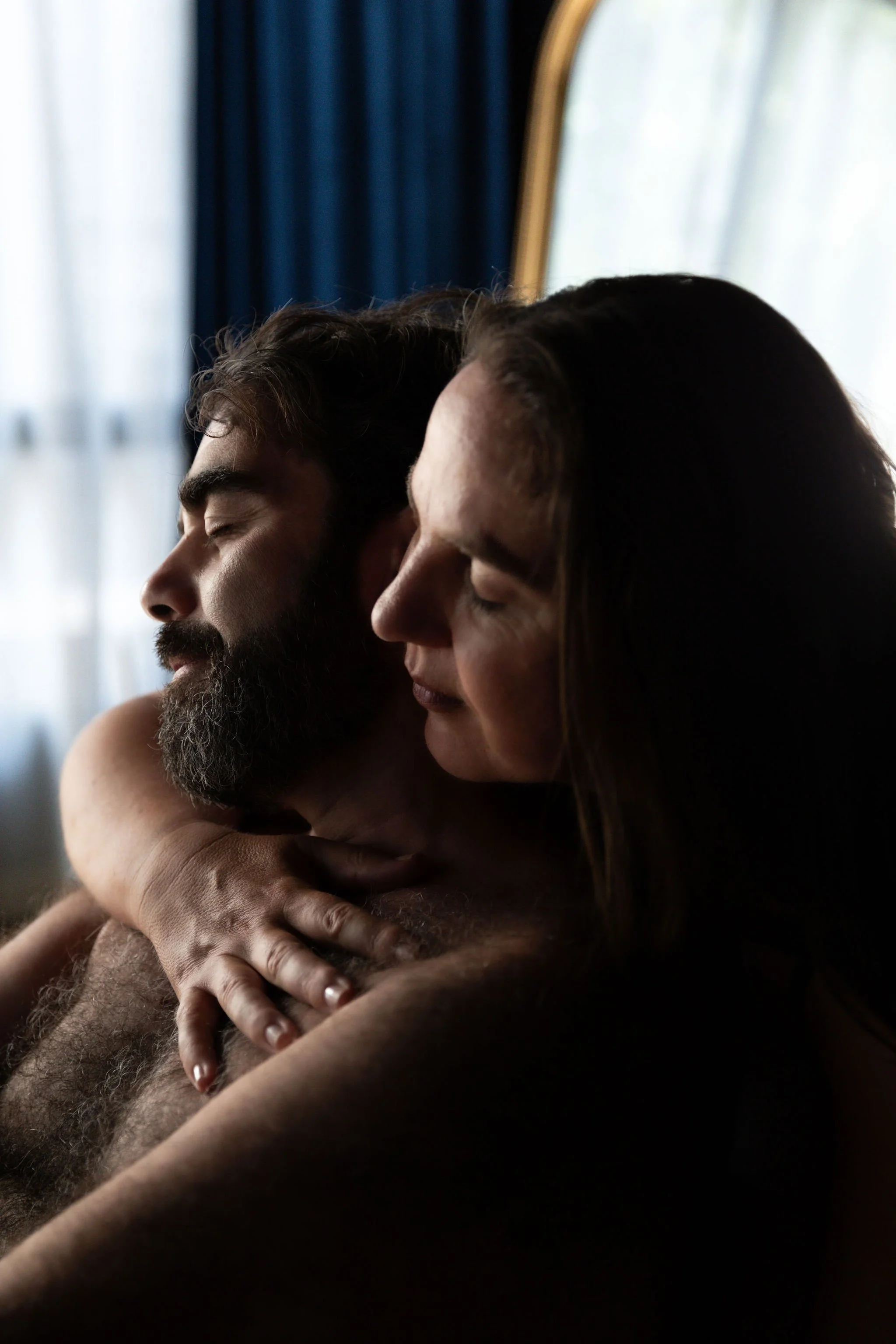 A man and a woman embrace closely, with eyes closed, in a intimate moment near a window with light curtains.