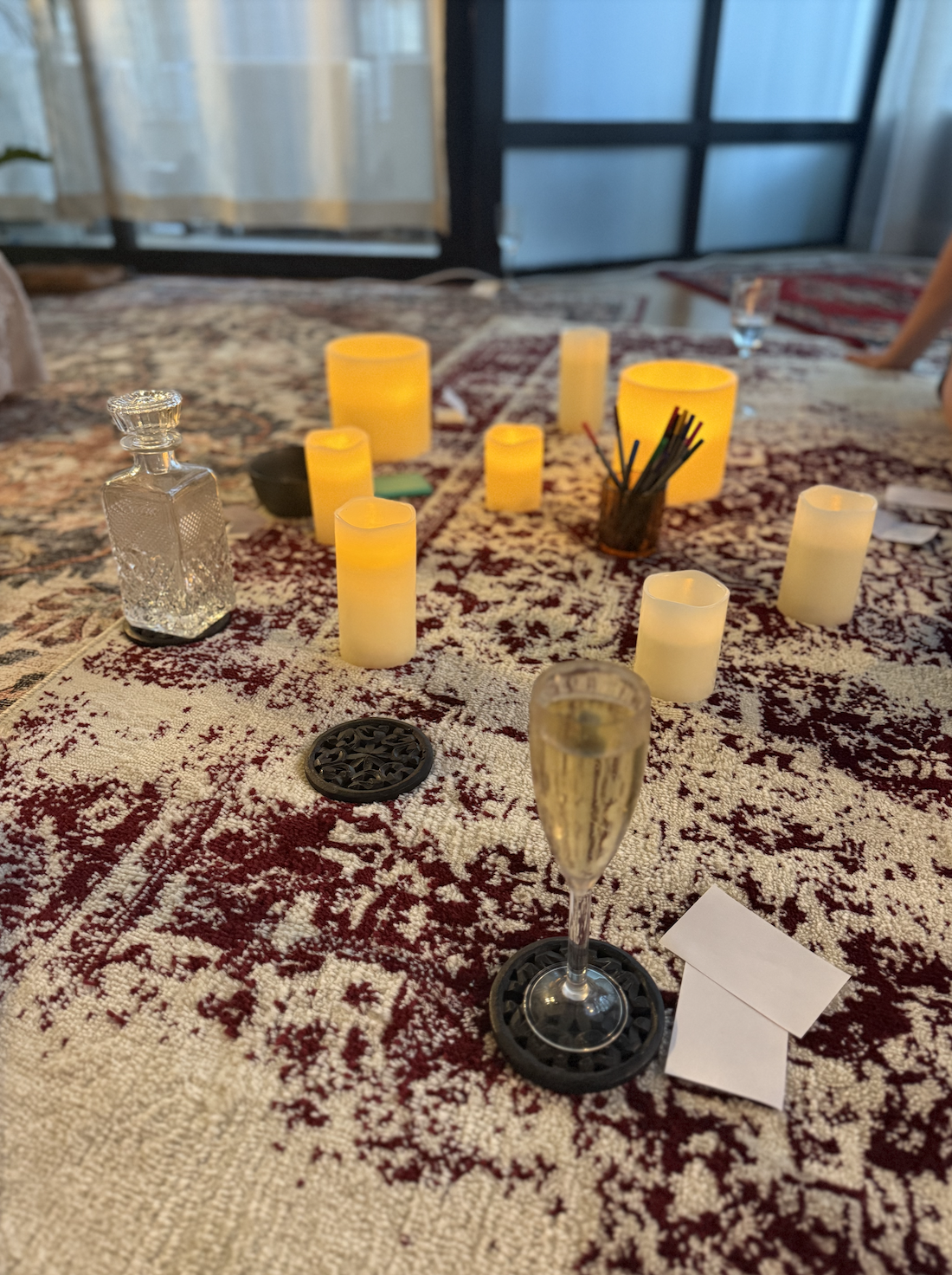 A table set with lit candles, a champagne flute, a glass decanter, and a small white card, with a patterned tablecloth in a cozy room.