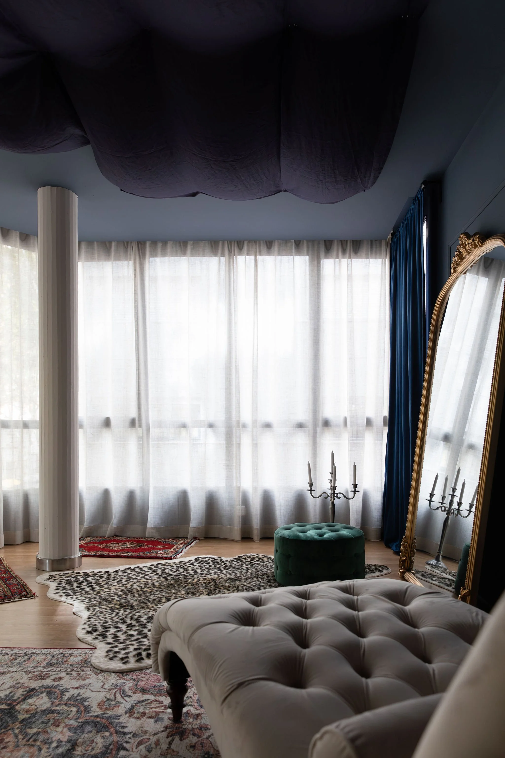 A room with large windows covered by sheer curtains, a white tufted chaise lounge in the foreground, a green ottoman, a full-length ornate mirror, a chandelier, and patterned rugs on a wooden floor.
