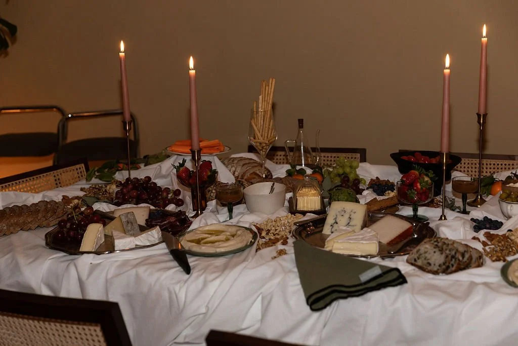 A long dining table set for a meal with various cheeses, grapes, bread, and fruit, decorated with lit pink candles.