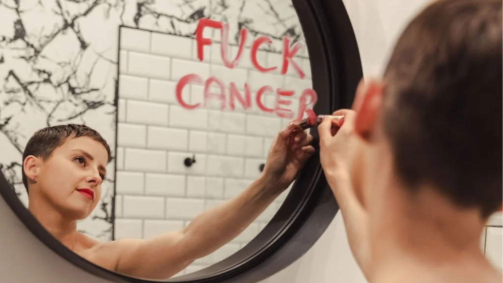 A woman is writing in red spray paint on a mirror, which reflects a white brick wall with the words 'F*CK CANCER' written on it.