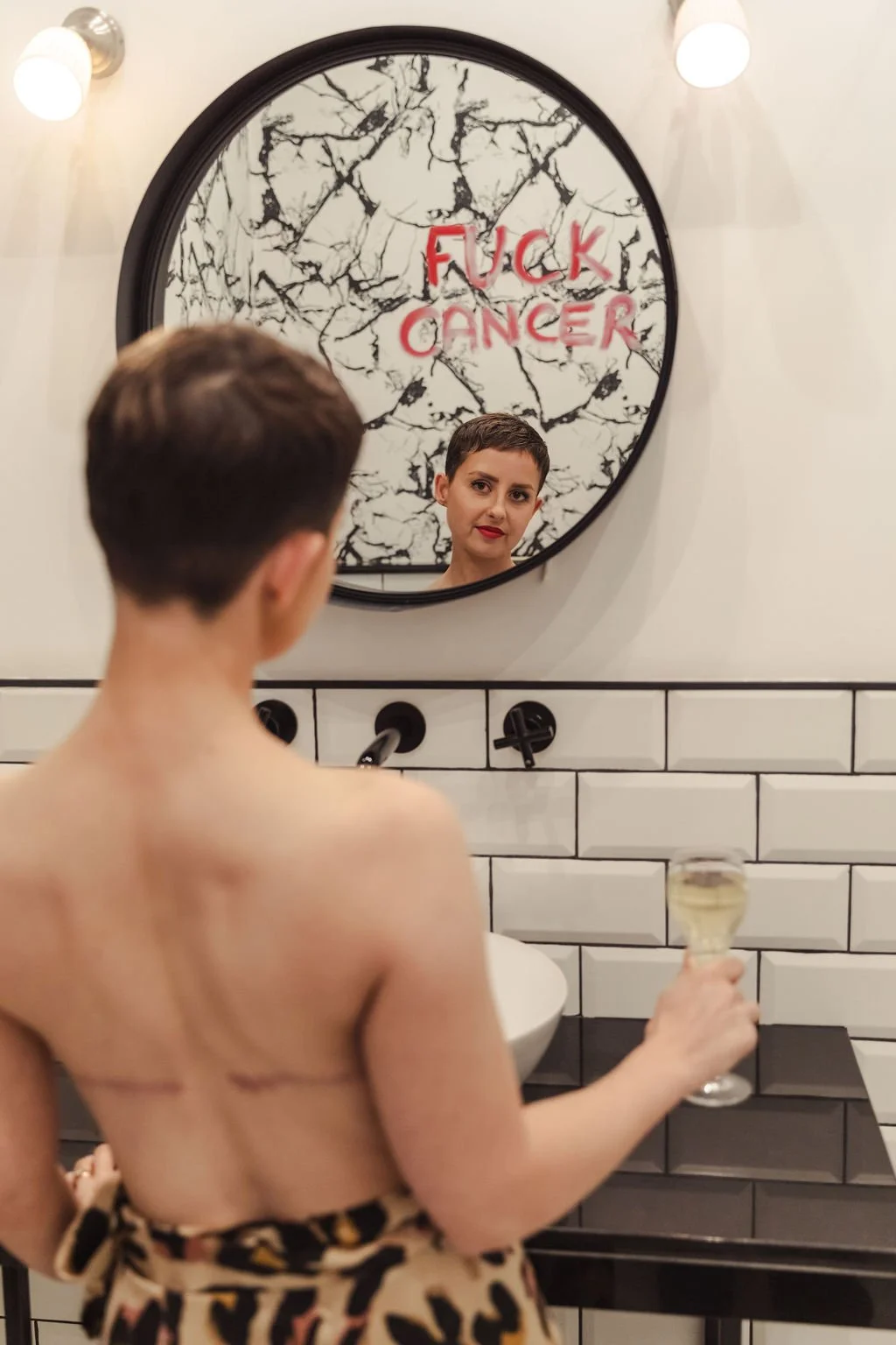 A woman with short dark hair and red lipstick looking at her reflection in a mirror. The mirror has red graffiti text that reads "FUCK CANCER" on a black and white cracked marble background. She is holding a glass of white wine in her right hand, and she appears to be topless with a leopard print towel tied around her waist.