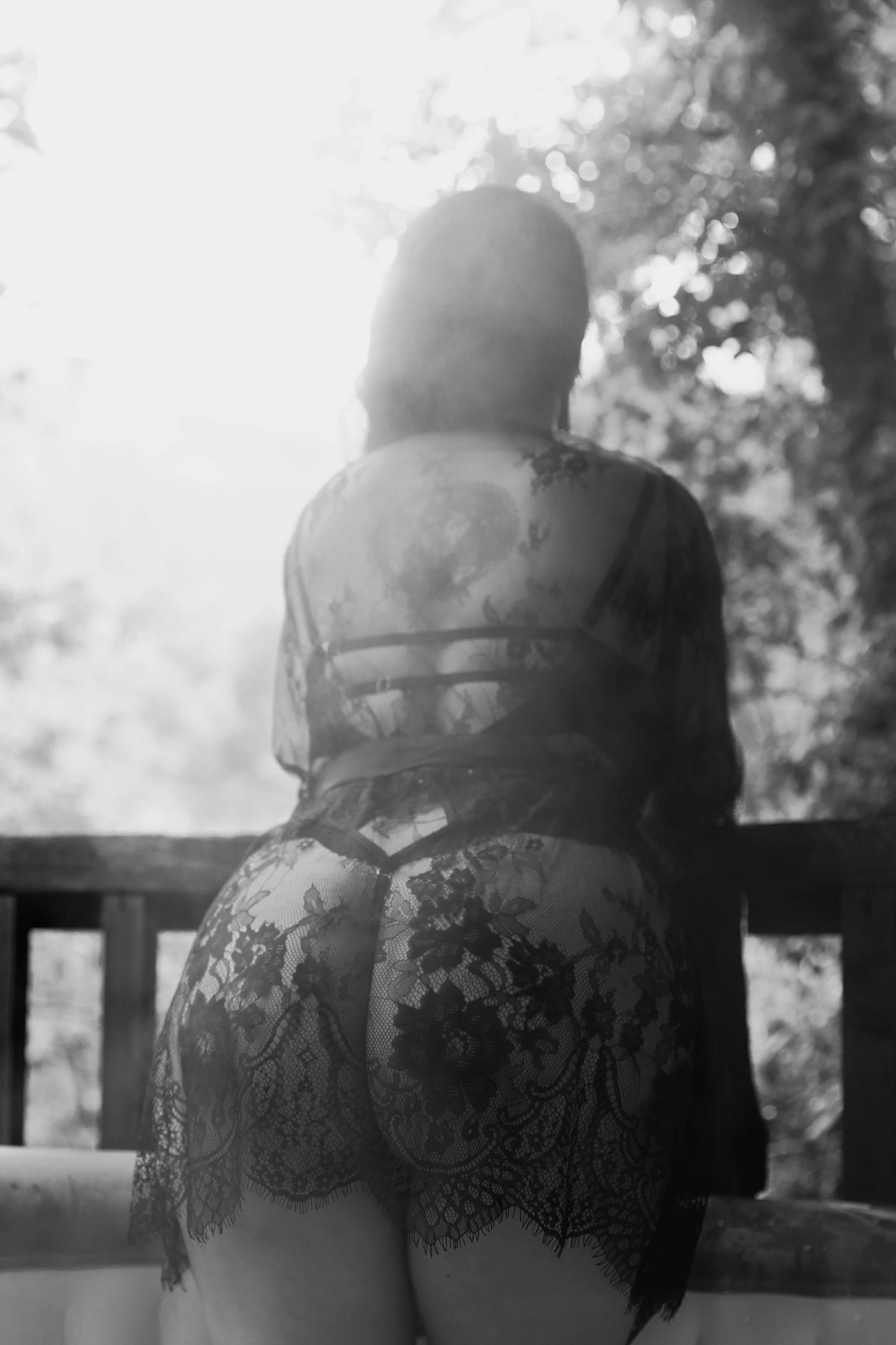 A woman in lace lingerie on a balcony, facing away, with sunlight and trees in the background.
