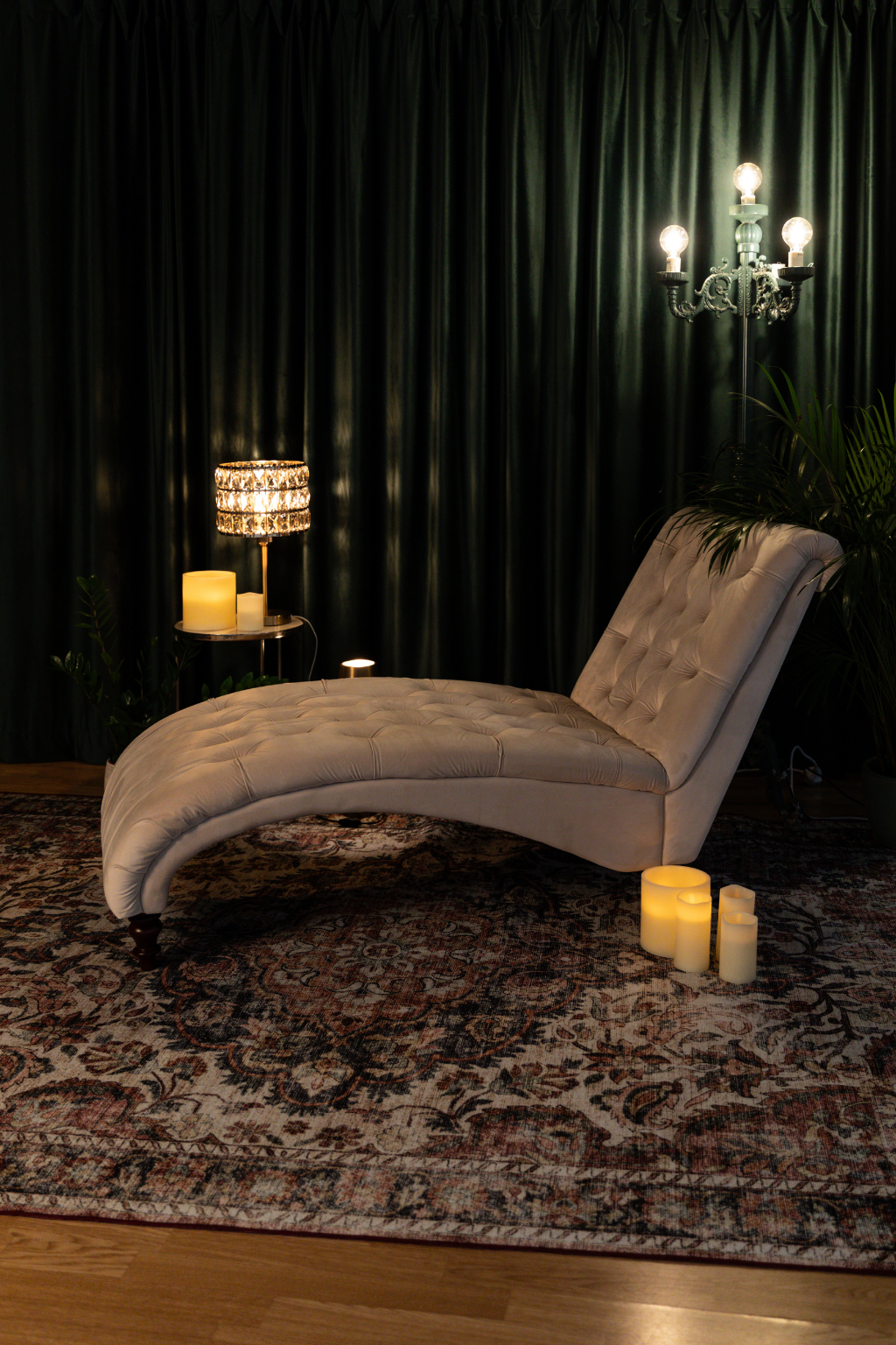 A cozy indoor scene with a beige chaise lounge, candles, a vintage chandelier, and a dark curtain backdrop.