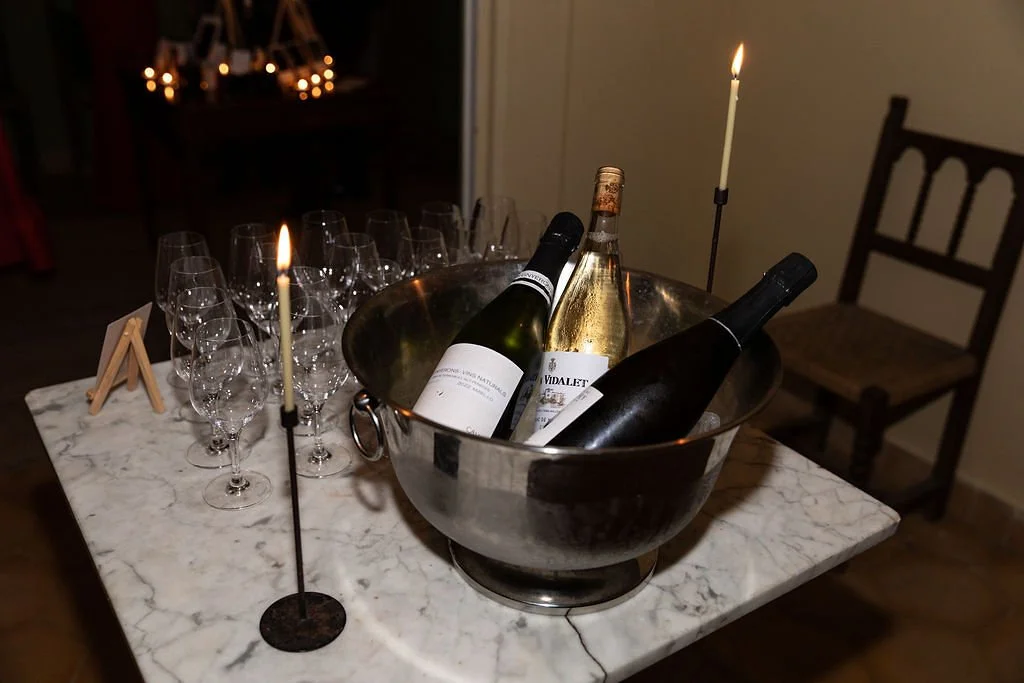 A marble table with empty wine glasses, a wine bucket containing three bottles of wine, and two lit taper candles.