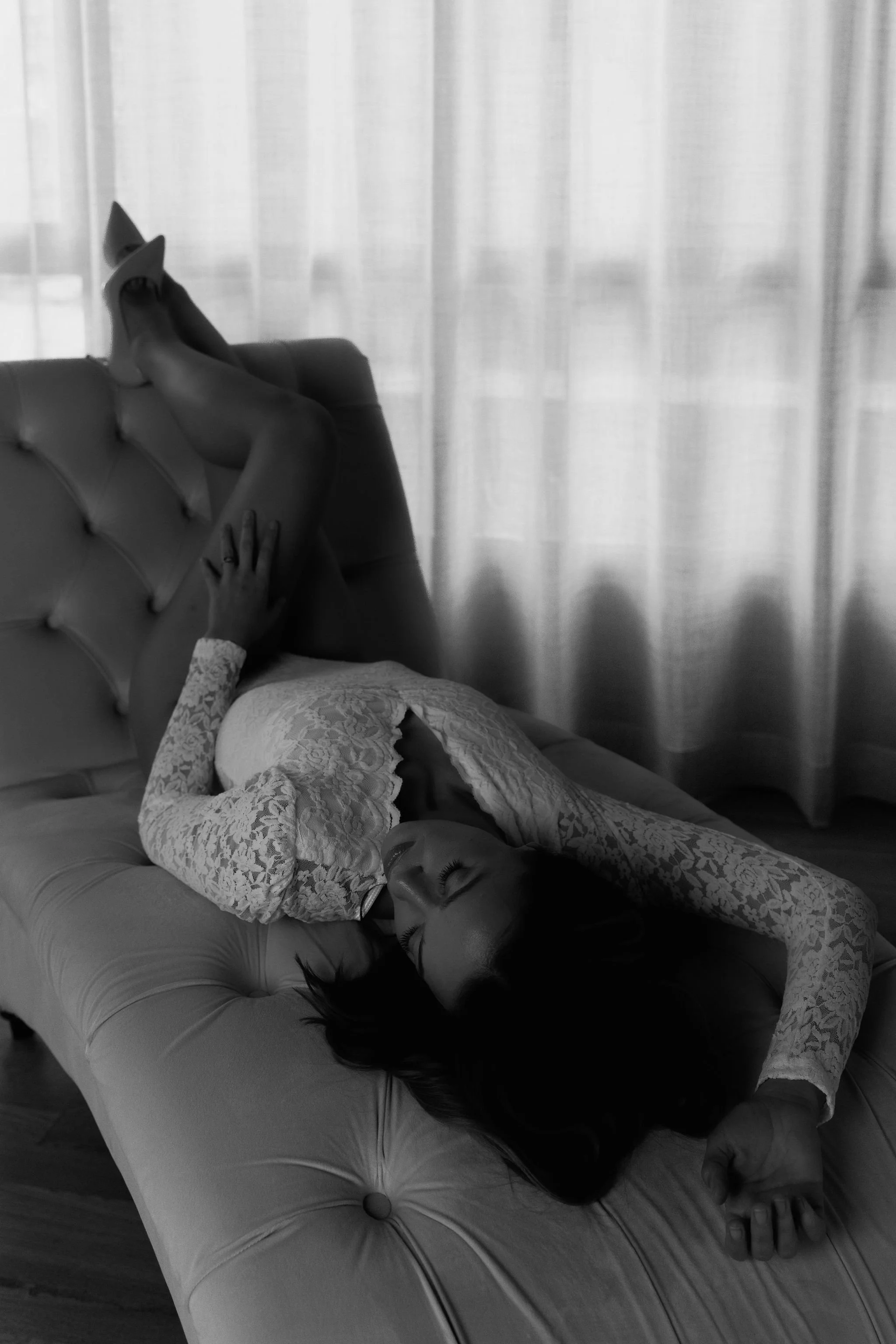 A woman with long dark hair, wearing lace long-sleeve top, reclining on a tufted chaise lounge, with her legs up on the back of the lounge, in front of sheer curtains.