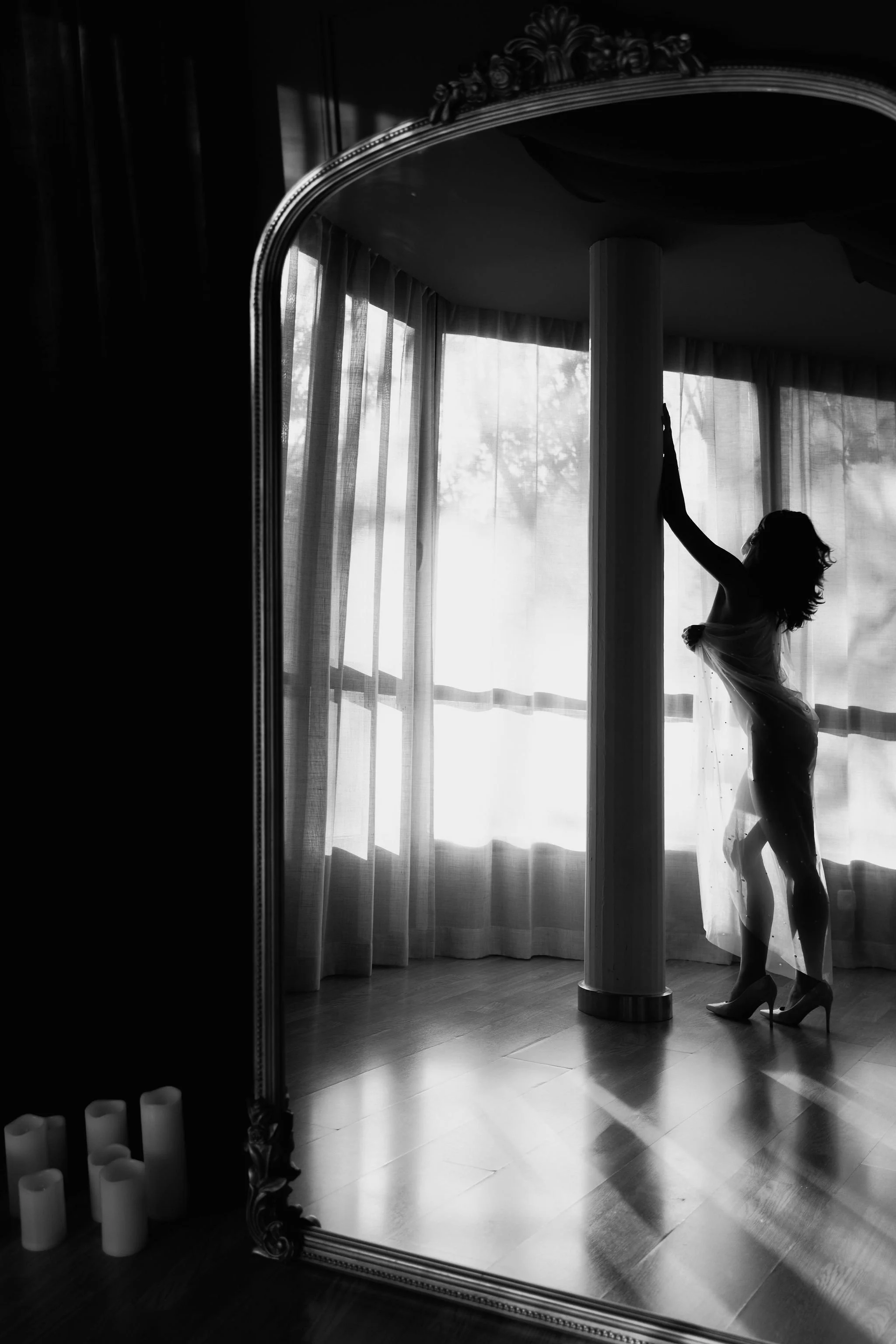 Silhouette of a woman in high heels standing by a large window with curtains, captured in black and white.