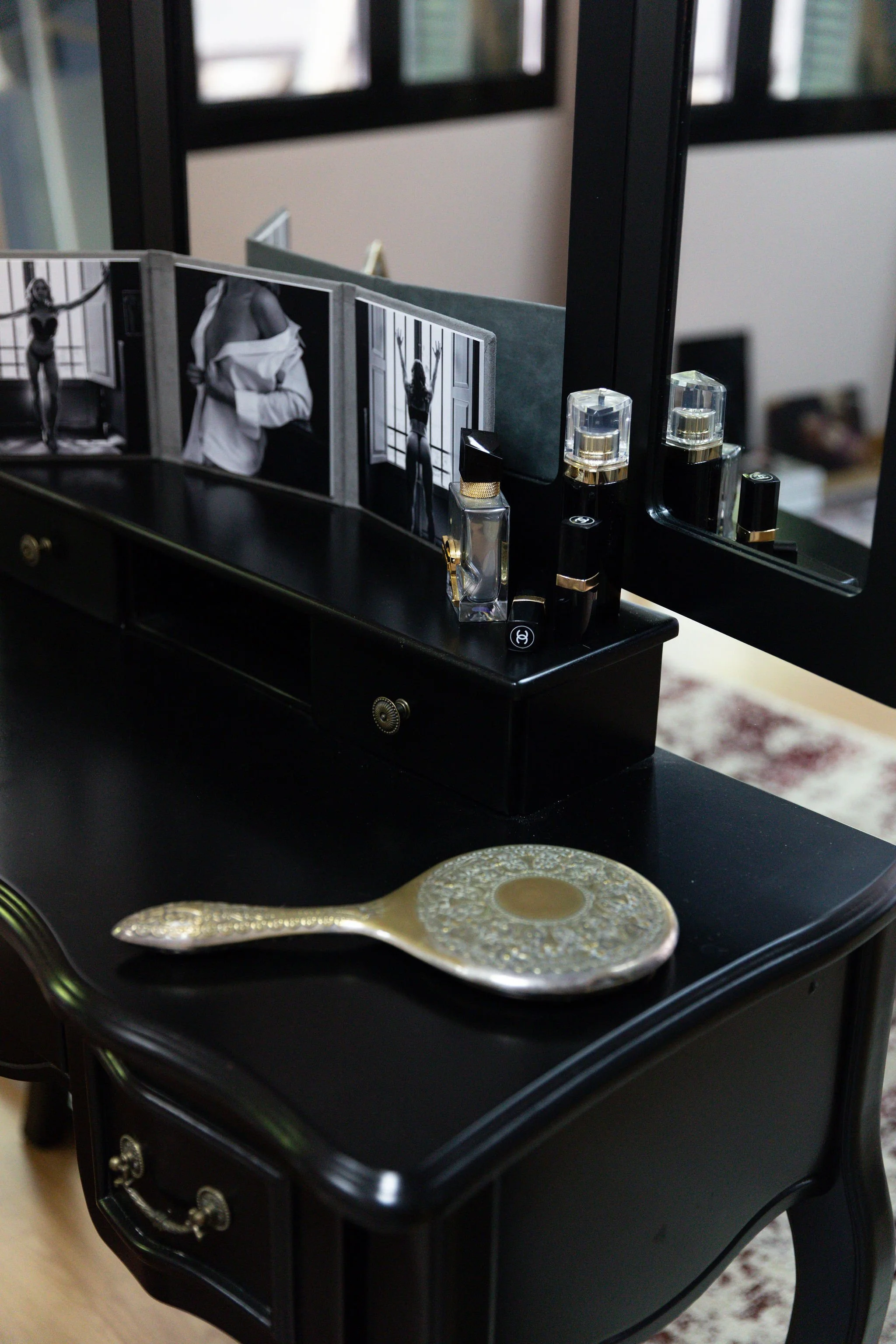 Black vanity table with perfume bottles, a mirror, black and white fashion photographs, and a decorative hand mirror, set in a room with large windows.