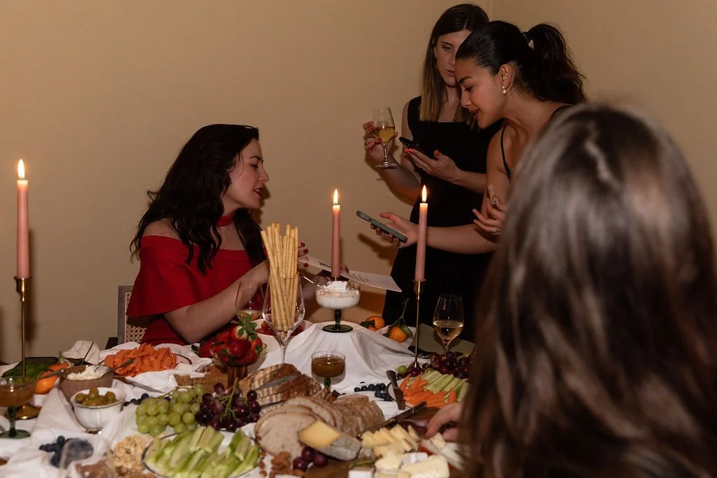 A celebrating woman in a red dress is sitting at a table set with food, candles, and drinks, while two women stand nearby, one with a glass of wine and the other looking at her phone, at a festive gathering.