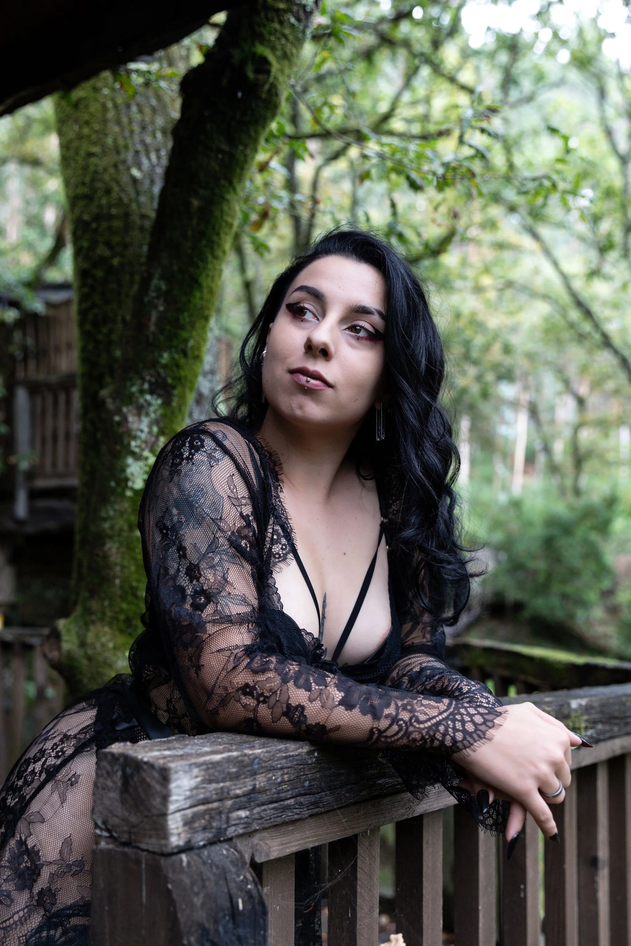A woman with black hair and makeup, wearing a black lace dress, leaning on a wooden railing outdoors in a wooded area.