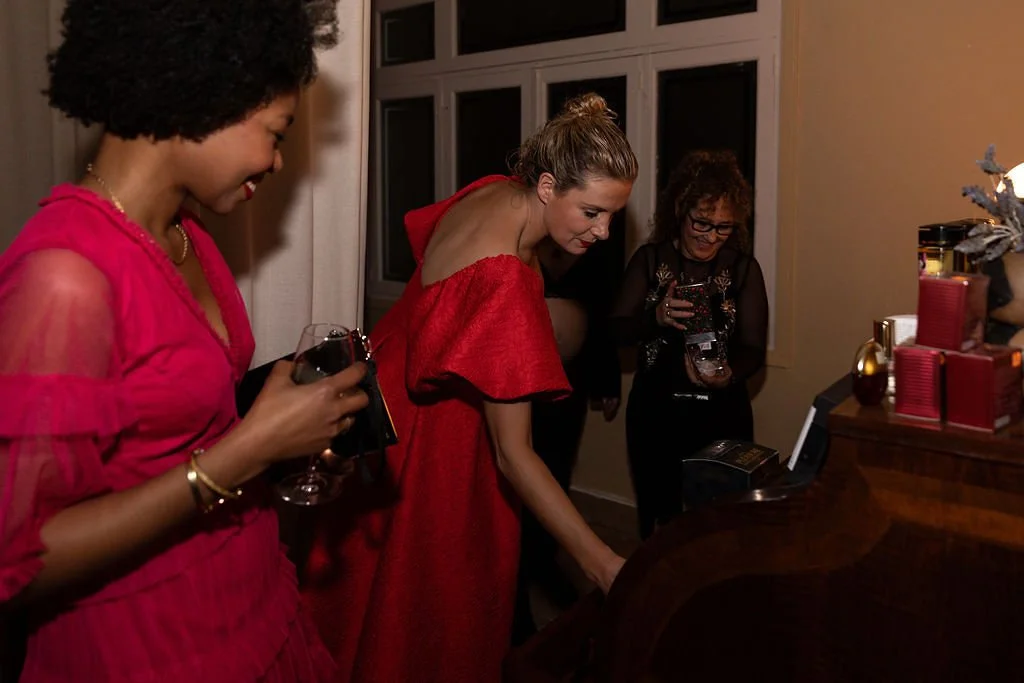 Three women in elegant dresses celebrating around a piano, with one woman playing the piano and others holding drinks, in a warmly lit room with windowpanes.