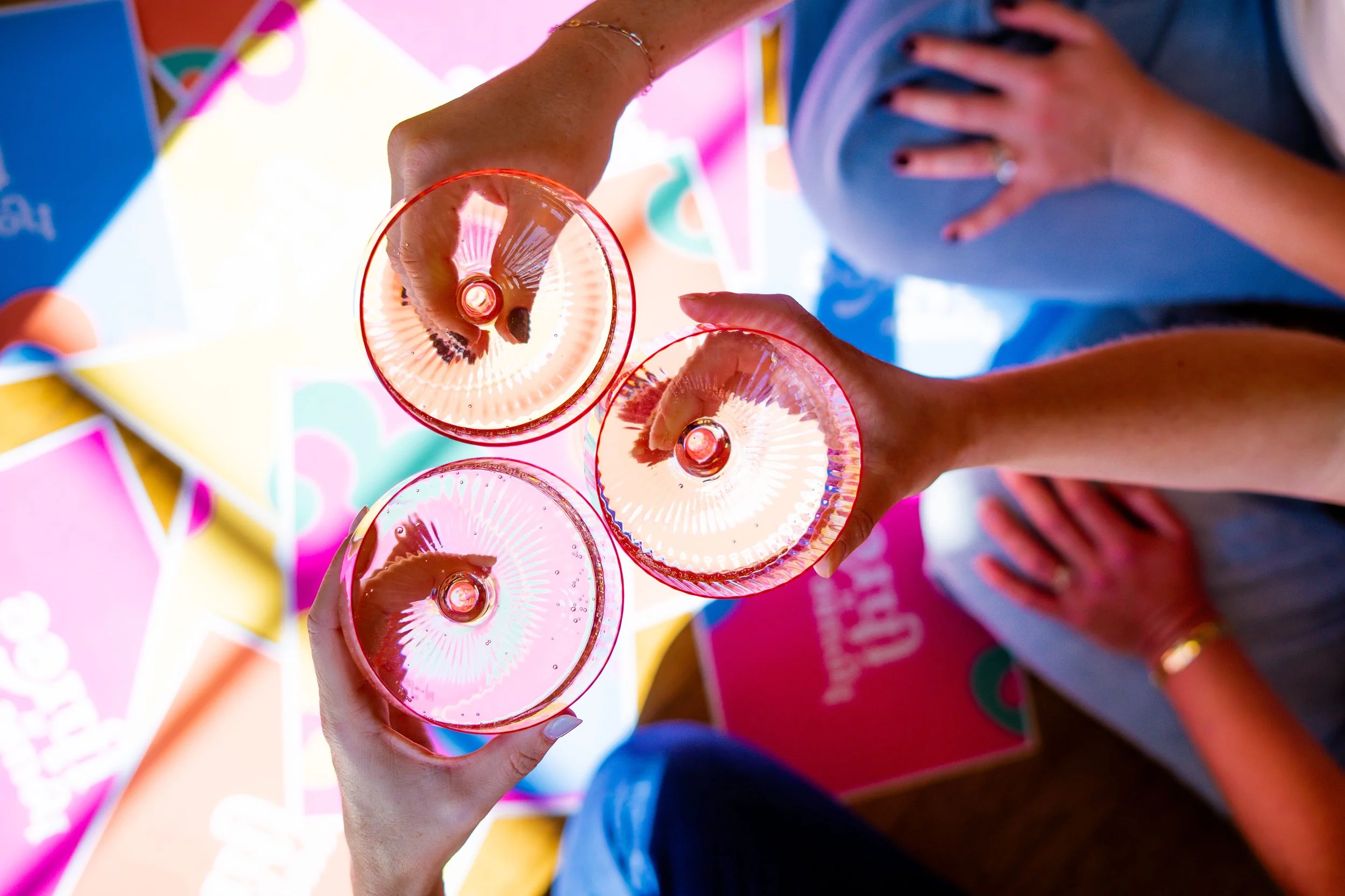 Three hands clinking pink drinks together over colorful prints in a celebratory, overhead view.