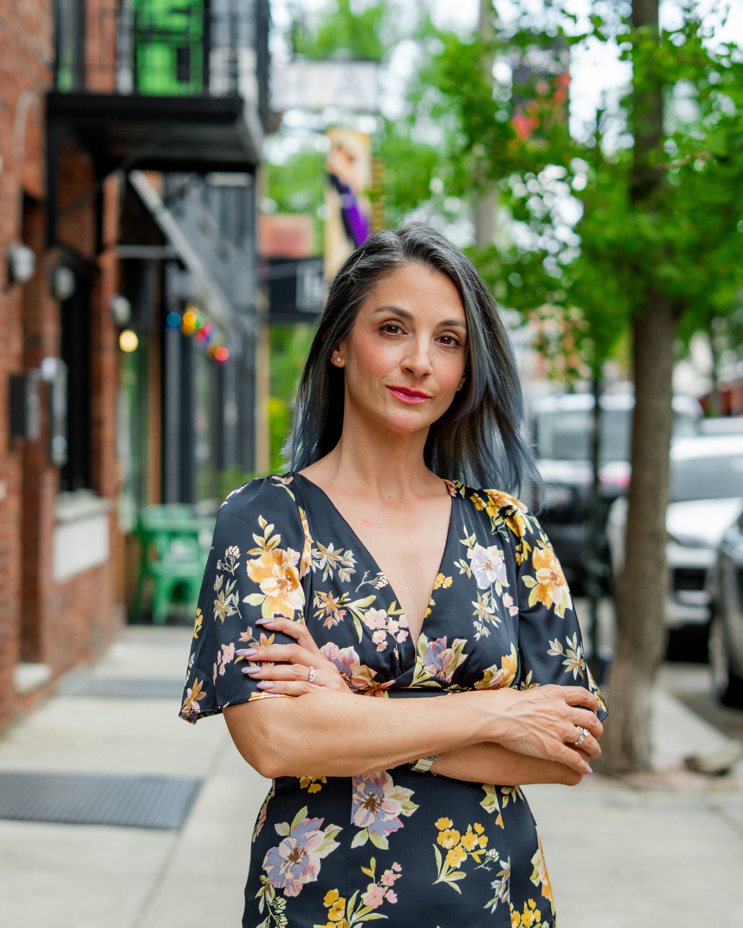 Capri Dessecker in a floral dress standing outdoors on a city sidewalk with trees and parked cars in the background.