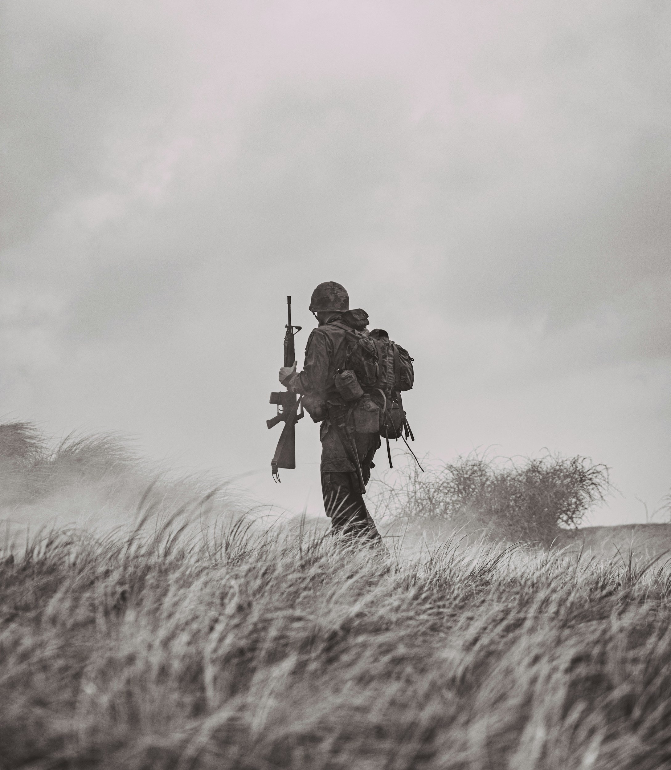A soldier dressed in camouflage gear walking through tall grass on a cloudy day, carrying a rifle and a backpack.