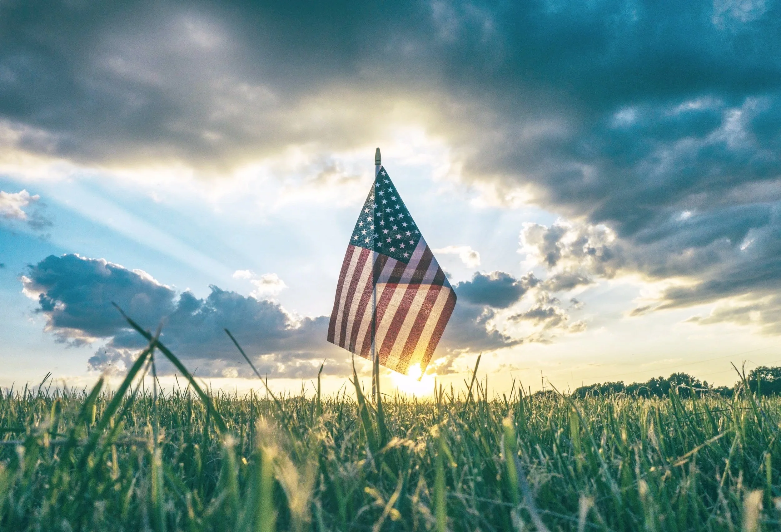 American flag waving in a grassy field at sunset with clouds and sunlight in the sky.
