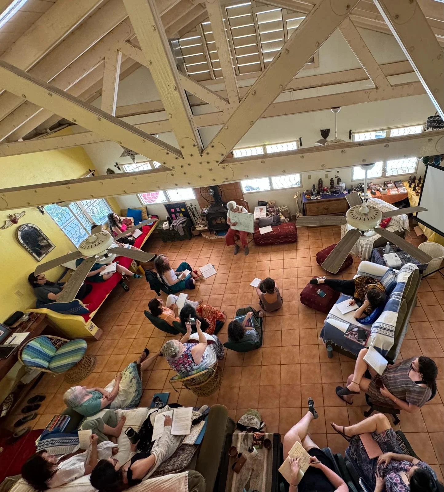 Indoor gathering of people sitting on sofas and chairs arranged in a circle, with a person teaching in the center. The room has large windows, a tiled floor, and ceiling fans. A wooden beam is visible at the top. Classrooms in Belize.