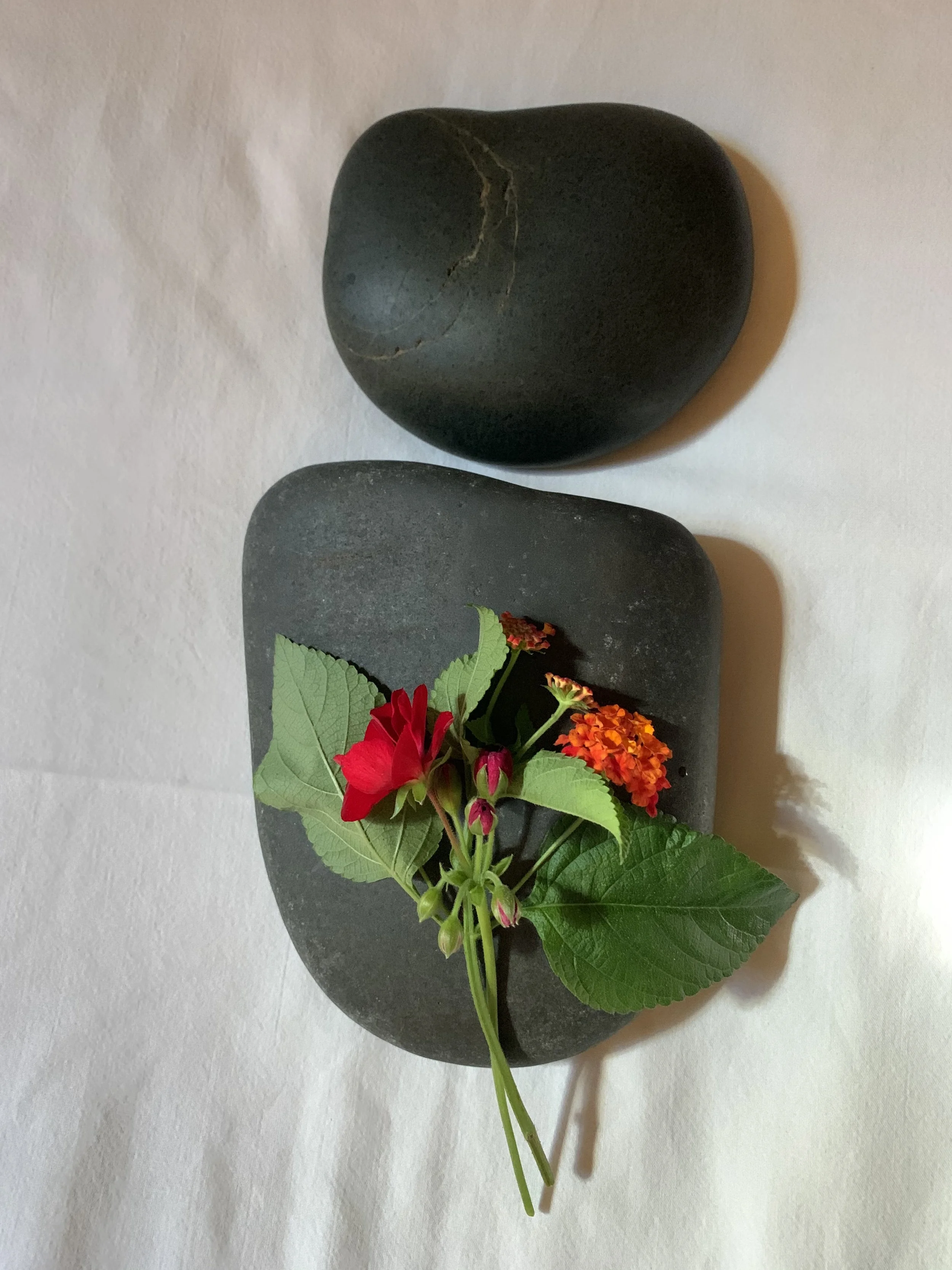 Decorative arrangement with geranium, lantana and, basil leaves, placed on a black basalt stone with a large black stone above.