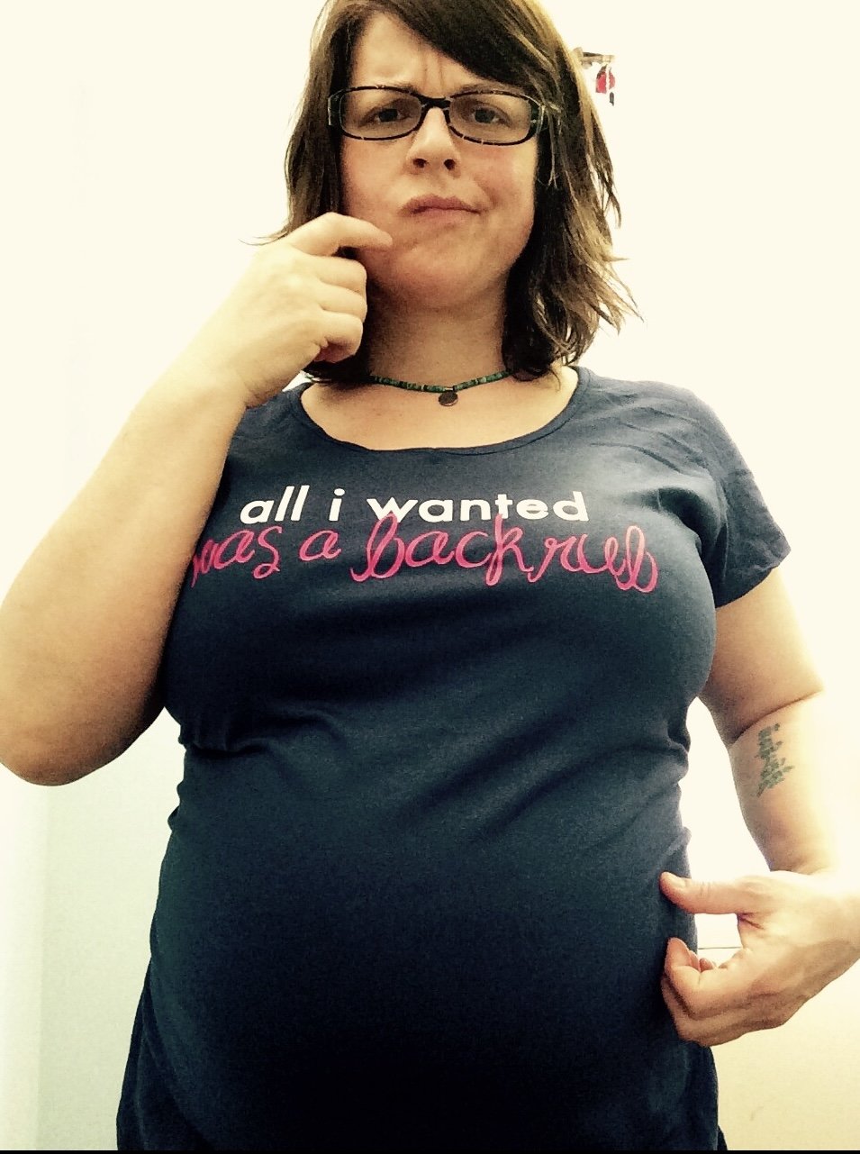 A confused looking pregnant woman with glasses and wavy brown hair, wearing a blue T-shirt that says 'all I wanted was a backrub' in pink and white text, standing against a plain background, with one hand on her hip and the other touching her chin.