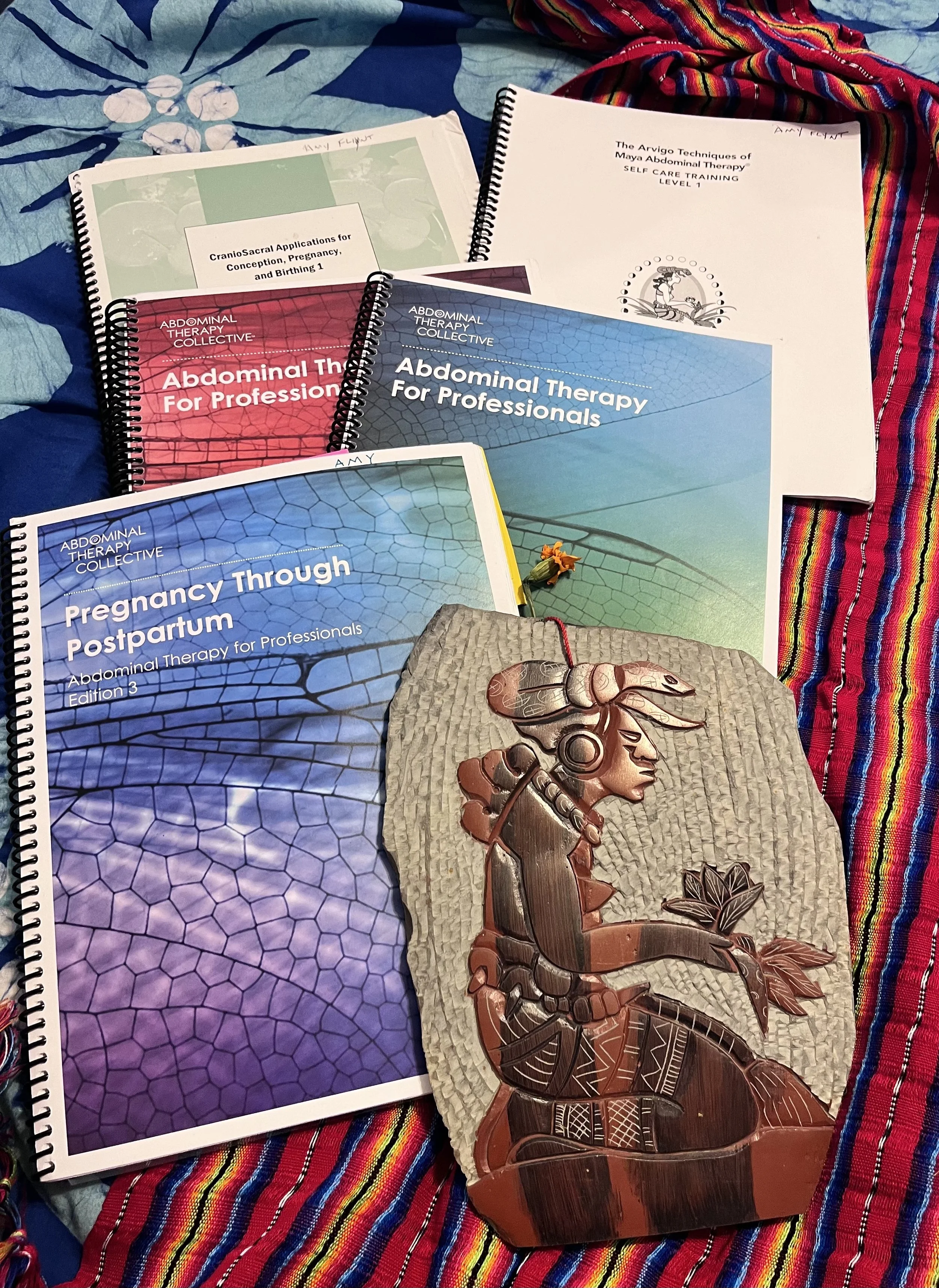 Collection of books on abdominal therapy, pregnancy postpartum, and self-care, along with a carved wooden plaque of a figure Ix Chel  holding a plant, all placed on a colorful textile background.