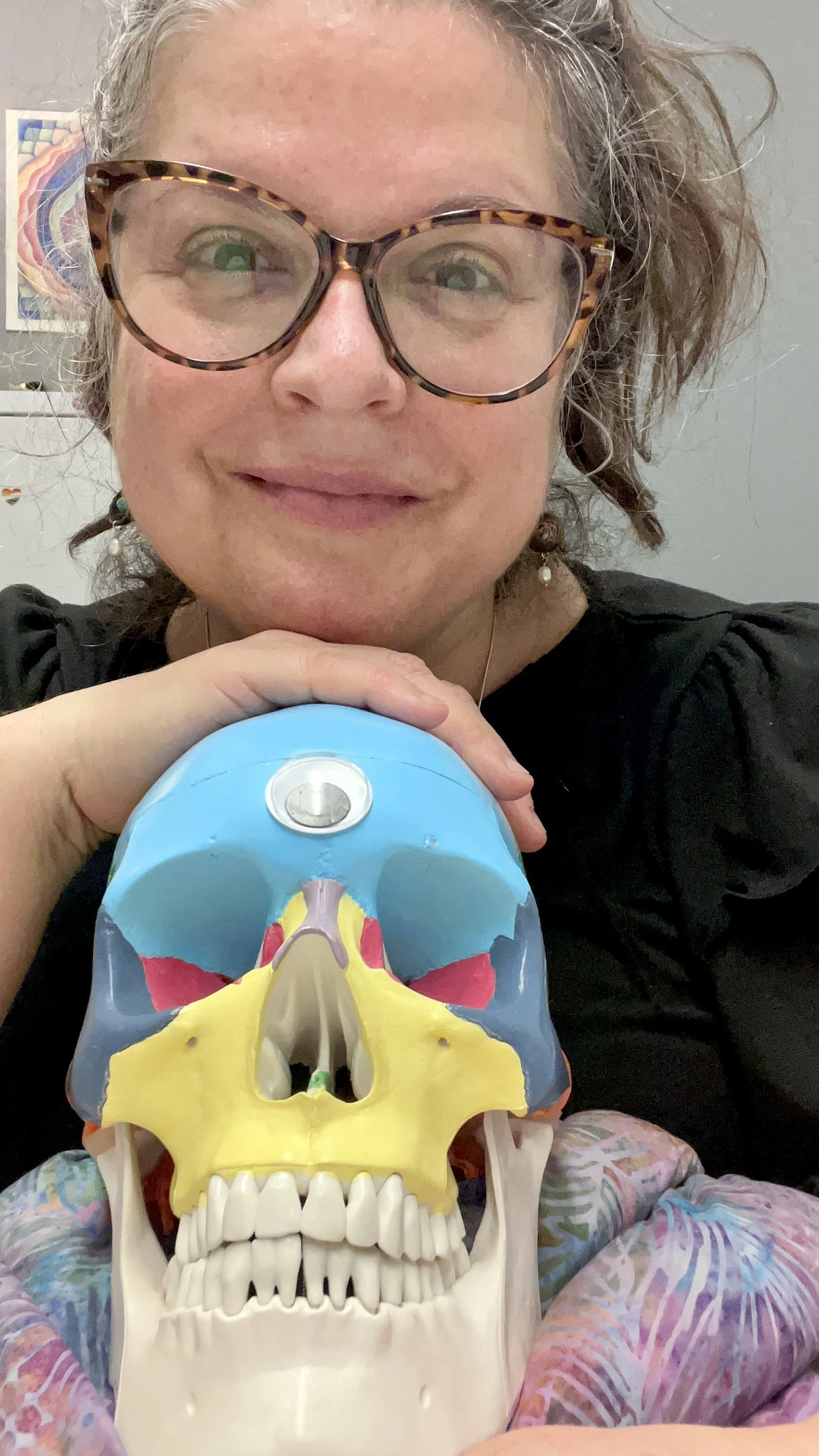 A woman with curly hair, wearing large tortoiseshell glasses, smiling, and holding a colorful anatomical skull model.