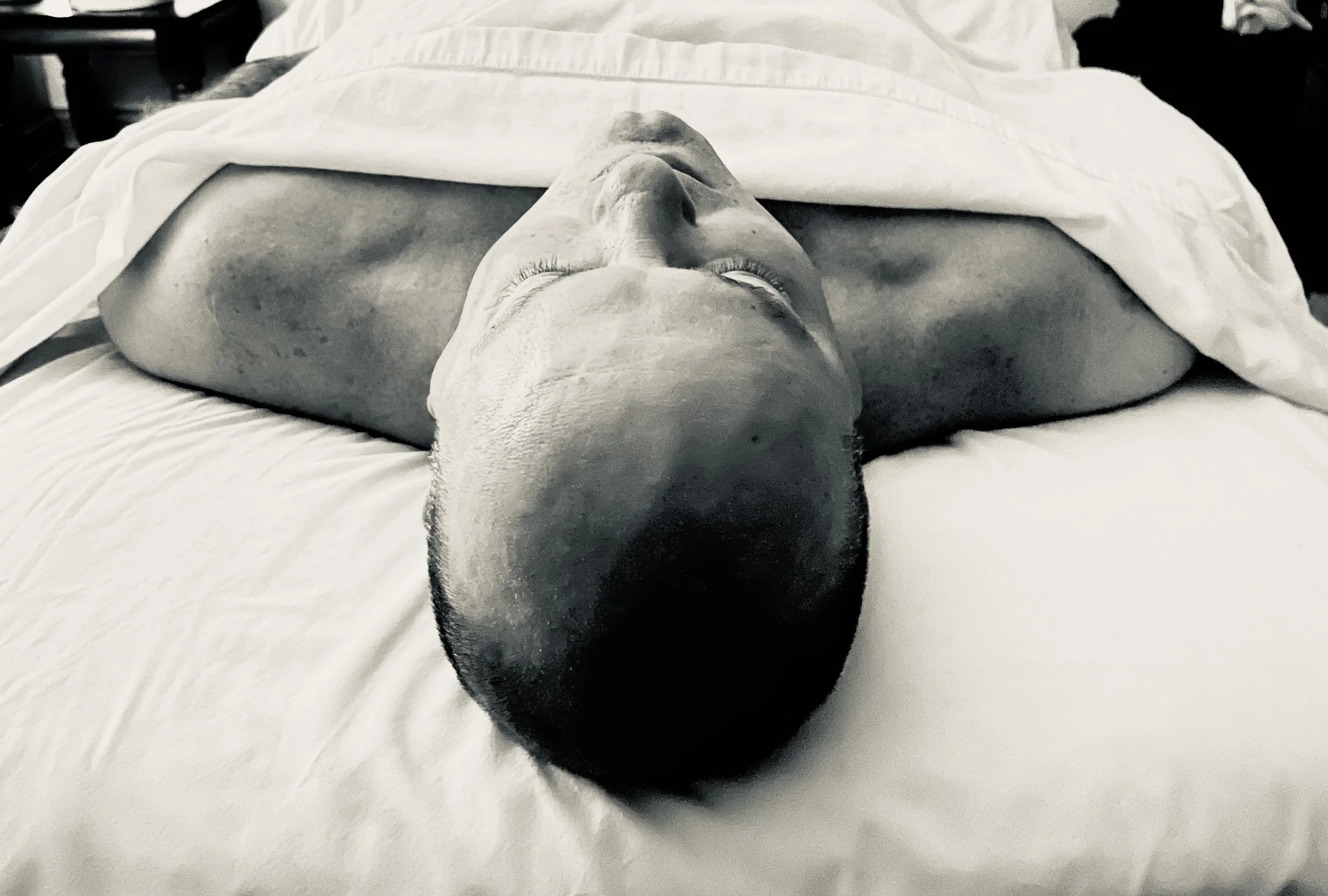Black and white photo of a person lying face up on massage table, draped with a white sheet up to their chest, with only their head and shoulders visible.