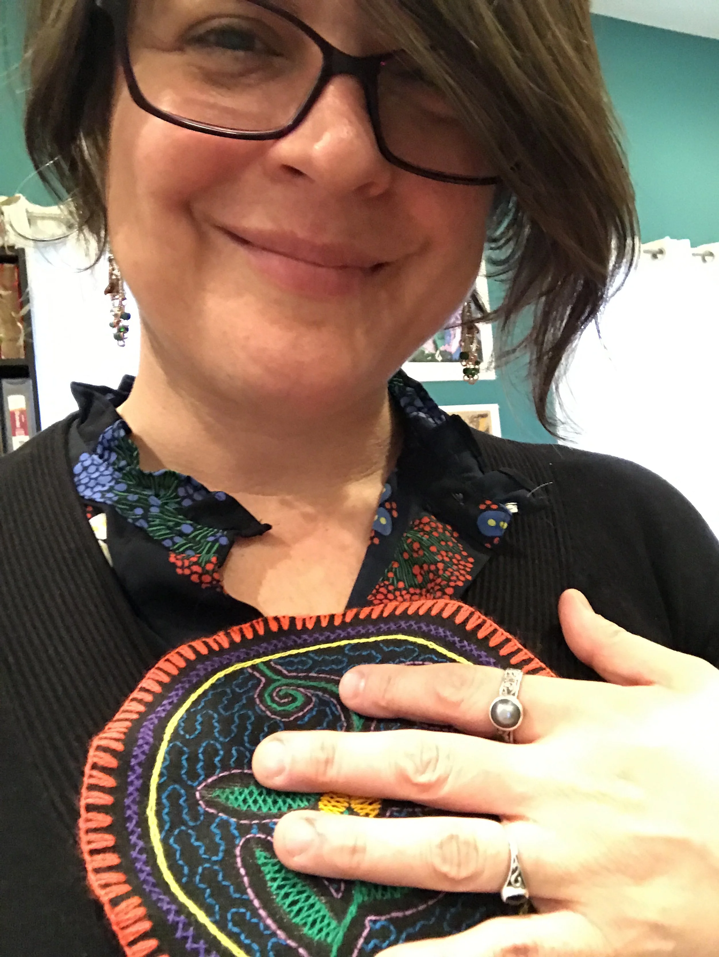 Close-up of a smiling woman wearing glasses, a black top with colorful embroidery, and a ring with a dark pearl, holding her hand over her chest.
