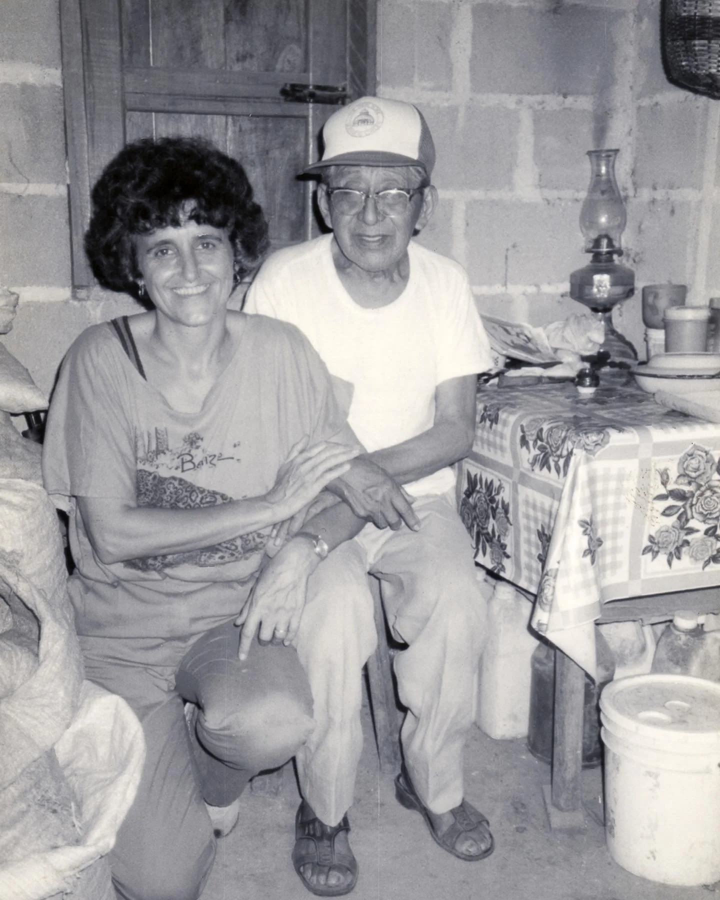 A black and white photo of an elderly man and a woman sitting together inside a rustic room. The woman has dark, curly hair and is smiling while holding the man's hand. Don Elijio Panti and Rosita Arvigo
