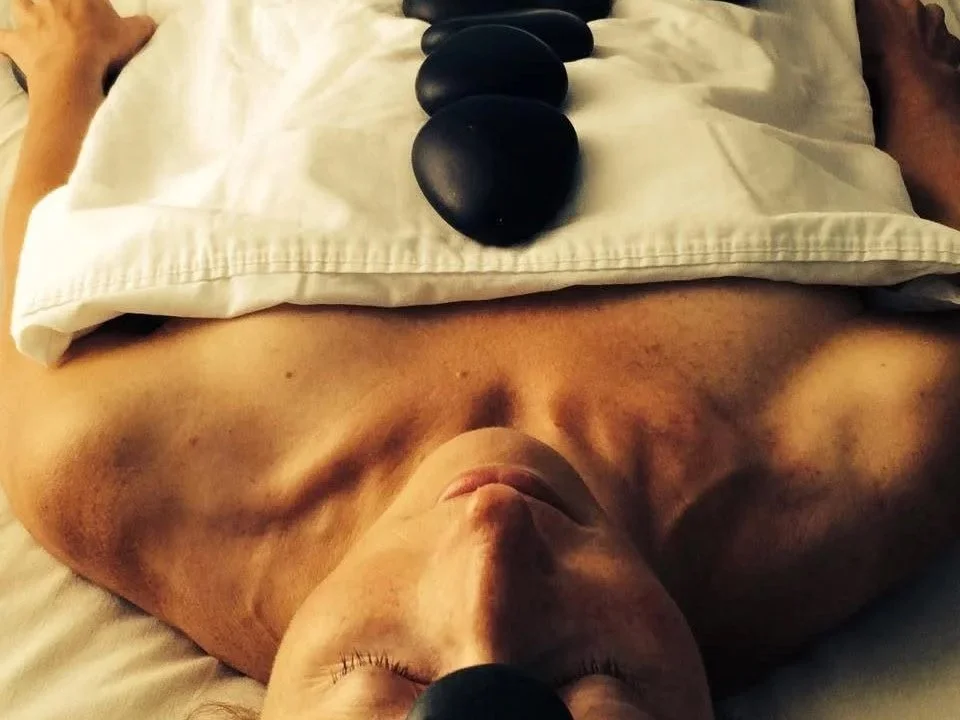 Person lying on their back with a massage stone on their chest and forehead, discreetly draped with a sheet..