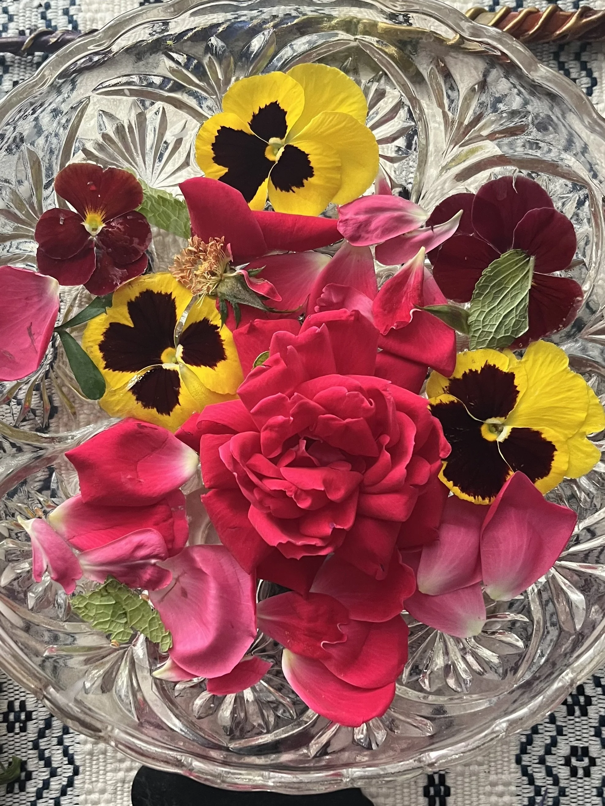 Glass bowl filled with colorful flower petals, including red roses, yellow and black pansies, pink petals, and green leaves.