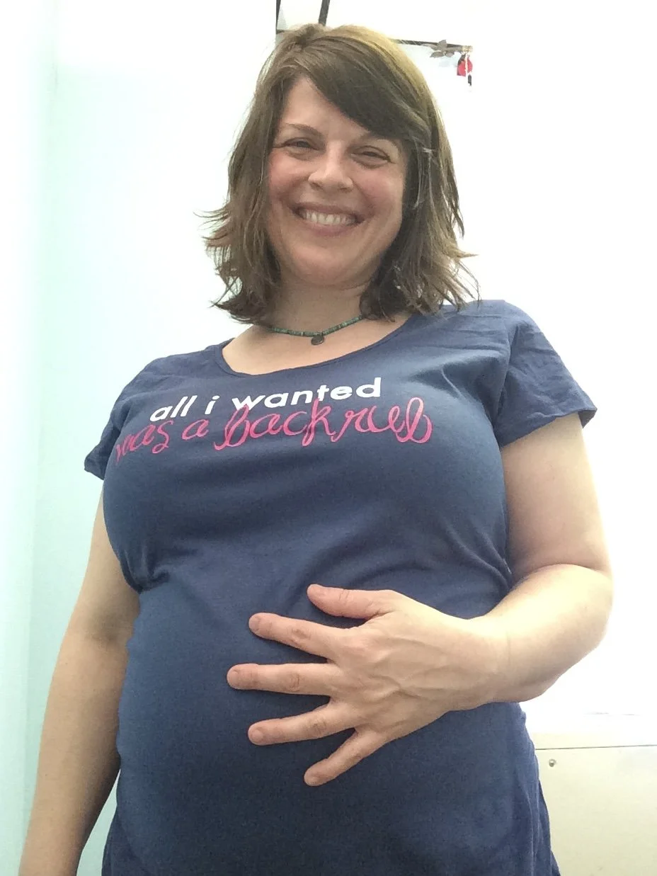 A woman smiling and holding her pregnant belly with her left hand. She is wearing a navy blue T-shirt with the text 'all i wanted was a backrub' in white and pink. She has shoulder-length brown hair and is wearing a necklace.