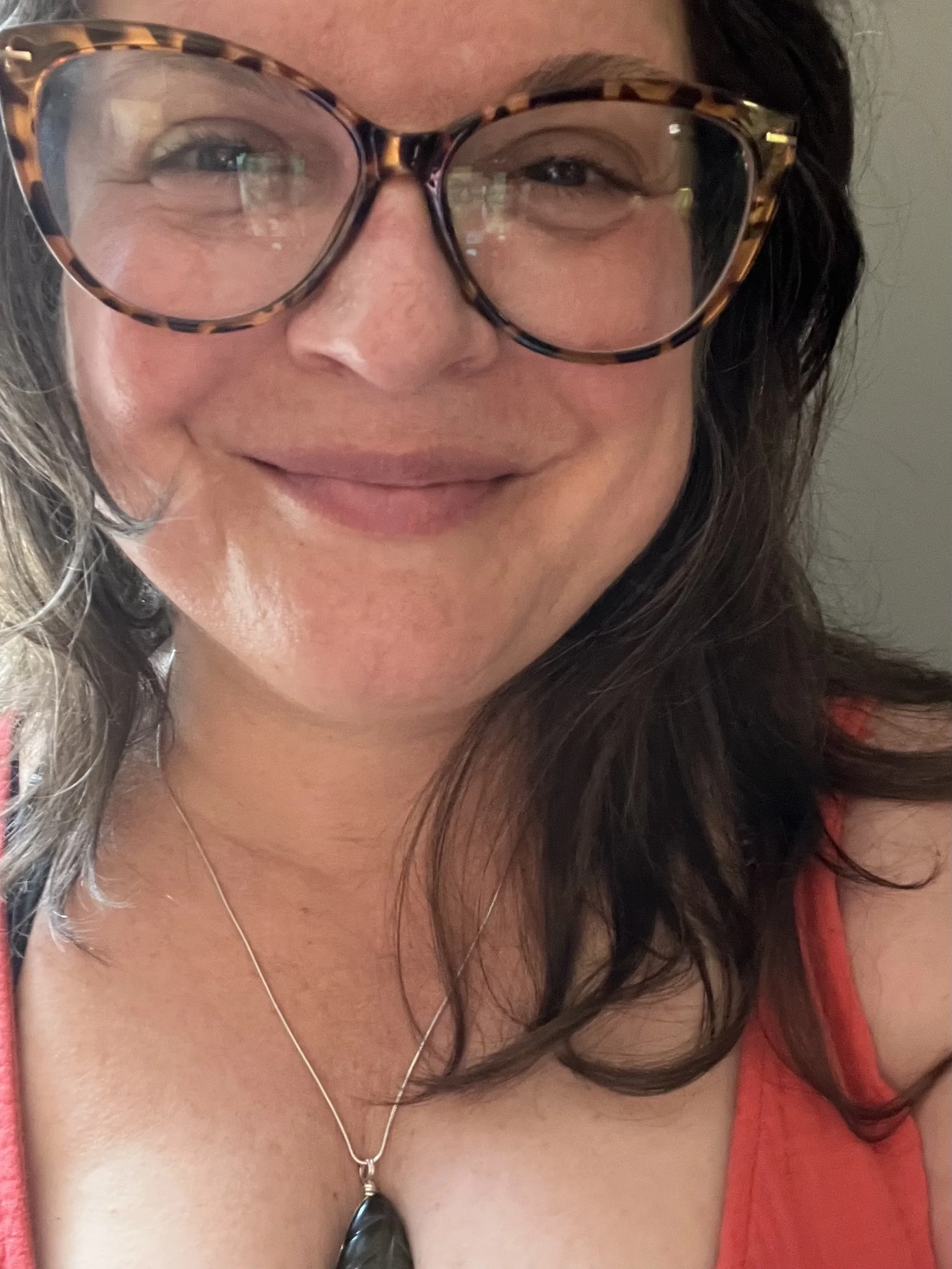 A woman with wavy dark hair wearing large tortoiseshell glasses, a coral sleeveless top, and a silver necklace with a black pendant, smiling at the camera.